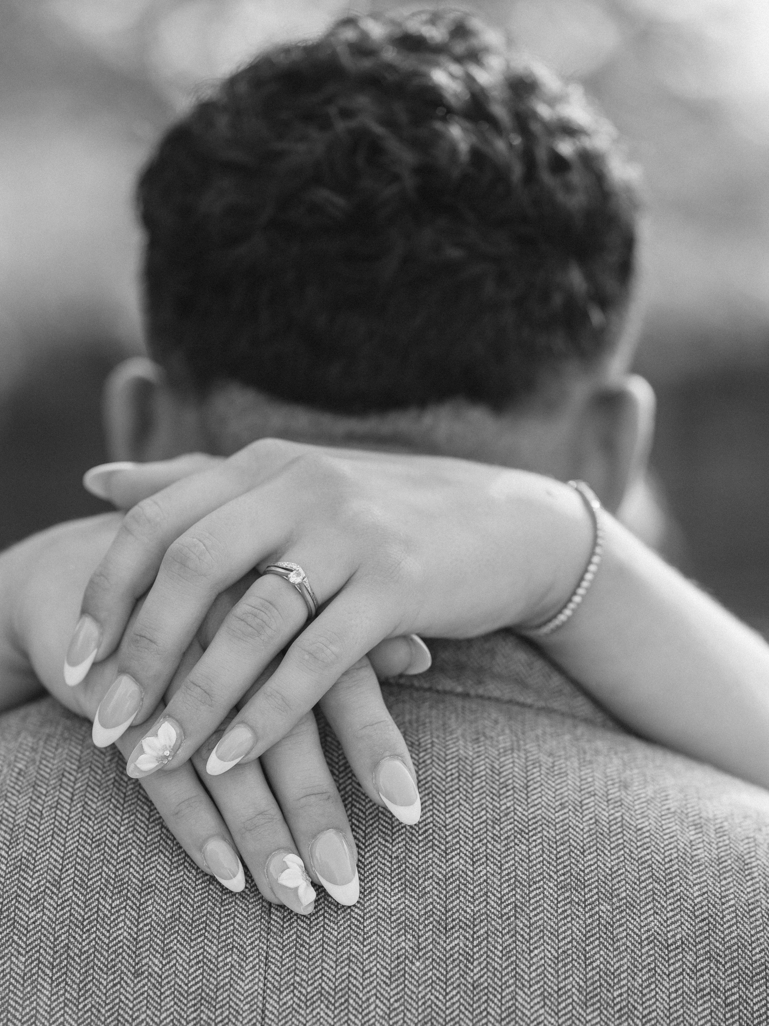 Close-up black and white photo of a person's back, with hands resting on their shoulders, one ring on the finger, wearing a necklace, and a textured fabric surface.