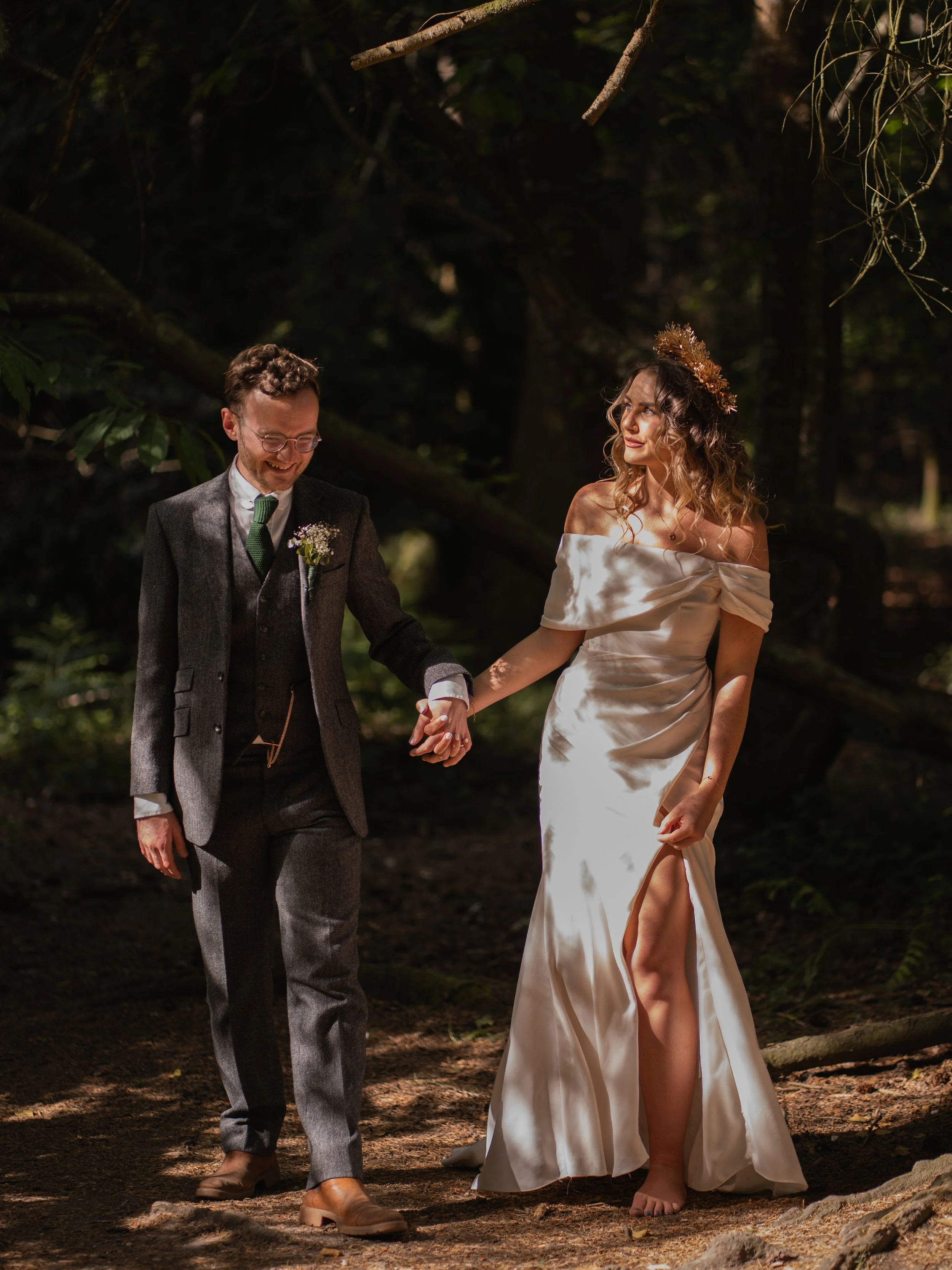 A man and woman holding hands in a forest, dressed in wedding attire. The man is wearing a suit with a boutonniere and glasses, smiling, while the woman is wearing a white off-shoulder wedding dress with a thigh-high slit, a floral crown, and is look
