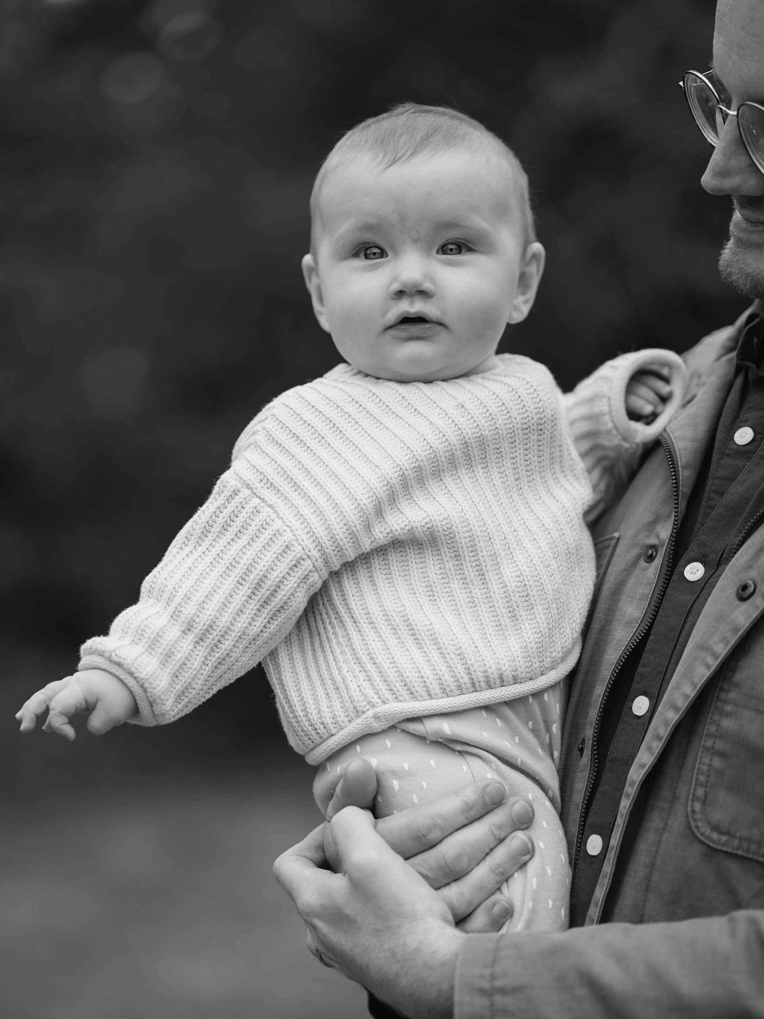 A black and white photo of a man holding a young child, possibly a toddler or baby, who is looking ahead with a curious expression. The man's face is partially visible, showing glasses and facial hair, and he is smiling. The child is dressed in a kni