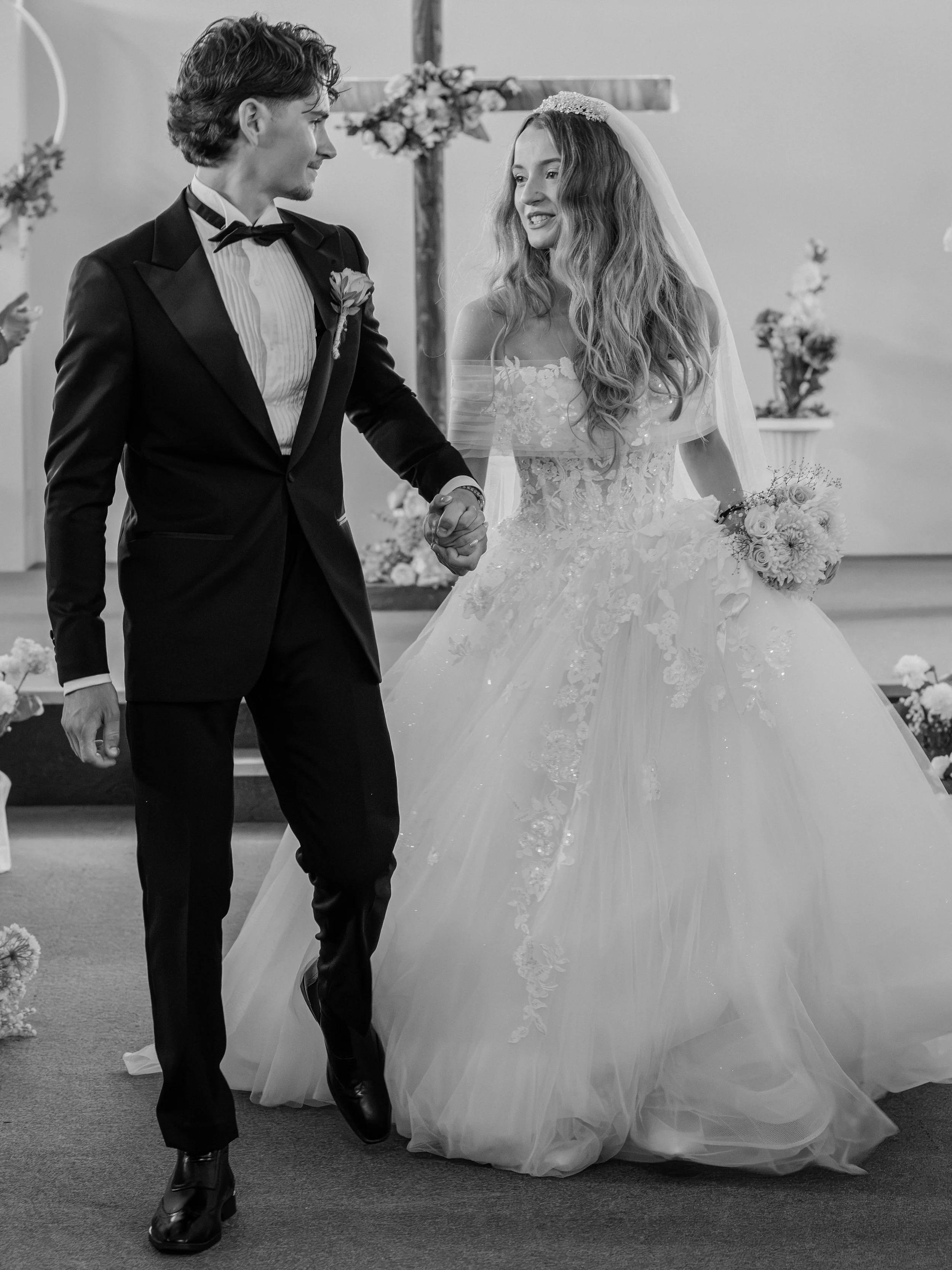 A black and white photo of a bride and groom holding hands during their wedding ceremony. The groom is dressed in a tuxedo with a bow tie, and the bride is wearing a wedding gown with lace details and holding a bouquet of flowers.
