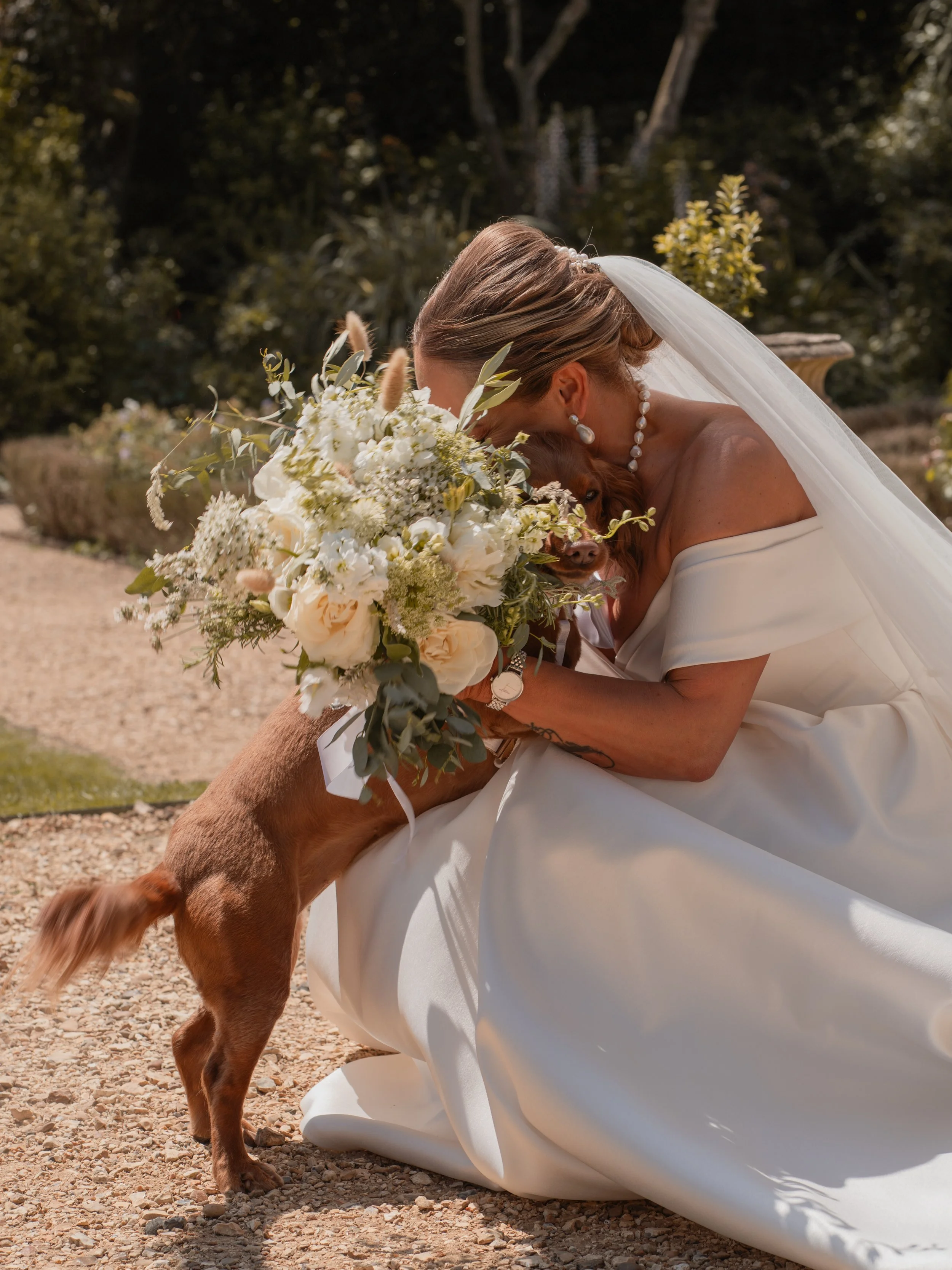 A bride in a white wedding dress and veil is hugging a golden retriever puppy, which is standing on its hind legs and licking her face, during an outdoor wedding ceremony with greenery in the background.