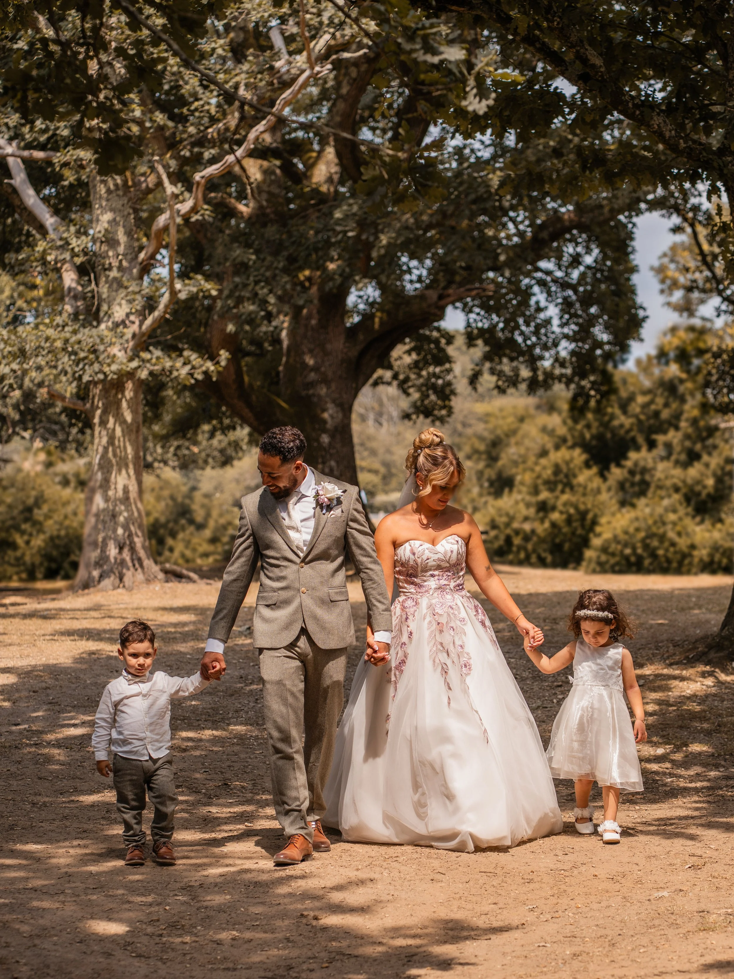 A wedding couple and three young children walking hand in hand outdoors under a large tree.