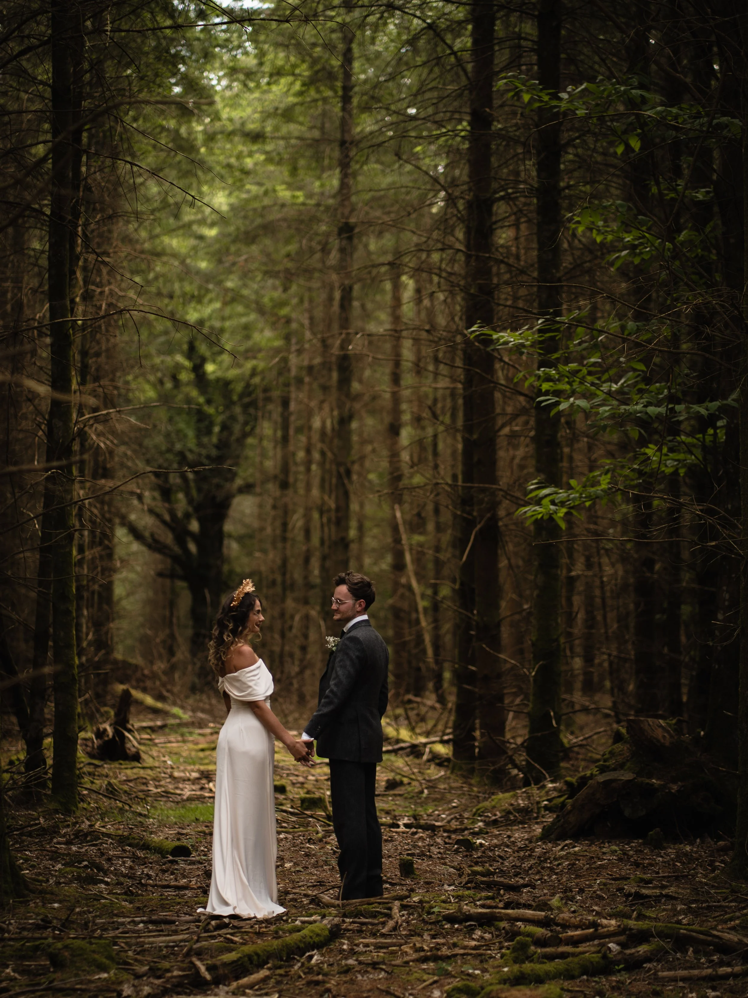 A bride and groom holding hands in a forest clearing, surrounded by tall trees and natural woodland scenery during daytime.