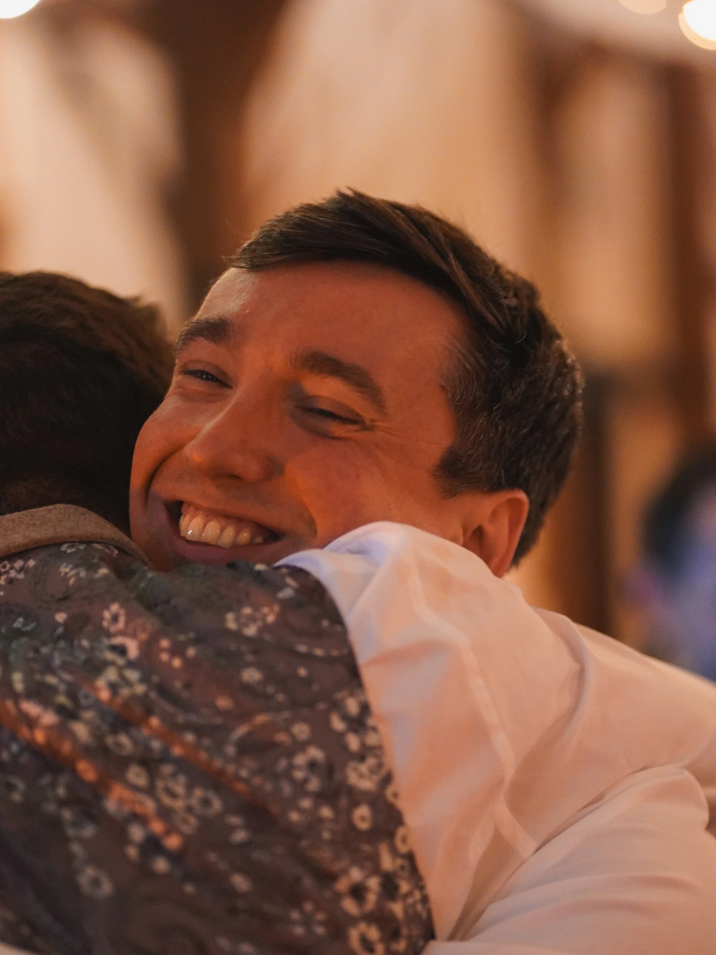 A man with a wide smile is hugging someone. The background is blurry with warm lighting.