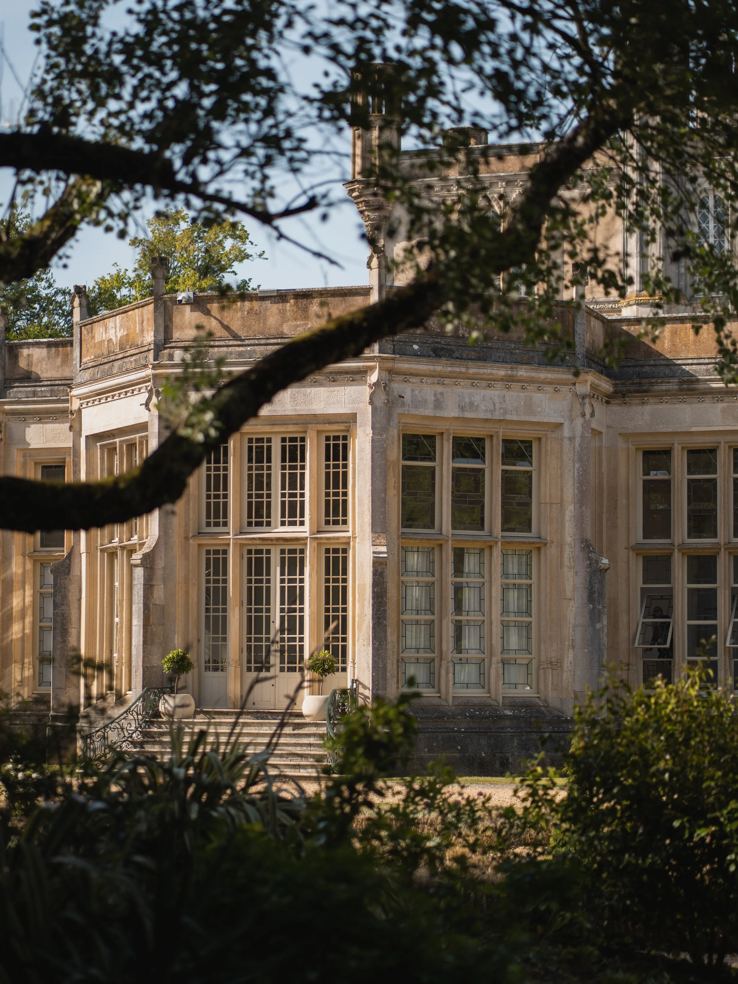 A historic building with large windows framed by stone, viewed through trees and foliage in the foreground.