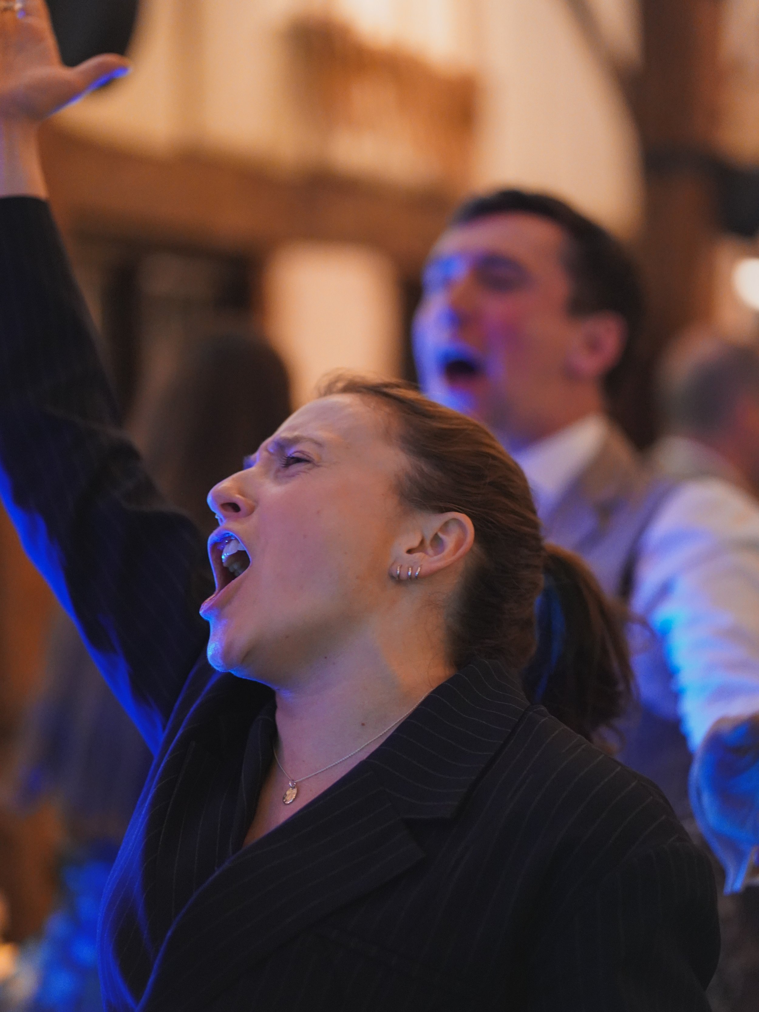 A woman with red hair and earrings is singing passionately with her eyes closed and mouth open, while a man behind her appears to be singing or speaking with his mouth open and eyes closed, in a dimly lit setting with blue lighting.