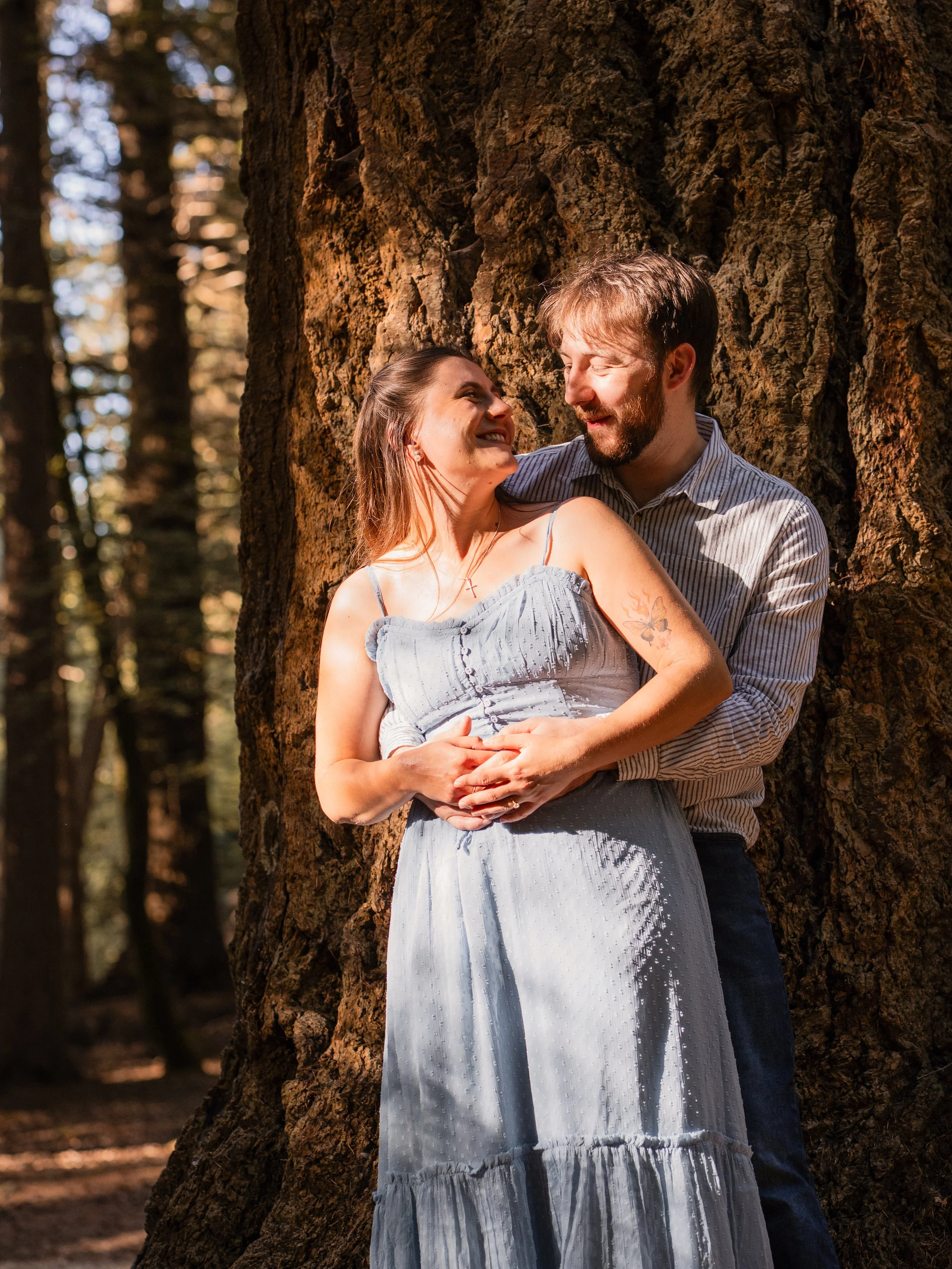A couple standing against a large tree trunk in a forest, sharing a joyful moment and looking at each other, with sunlight filtering through the trees.