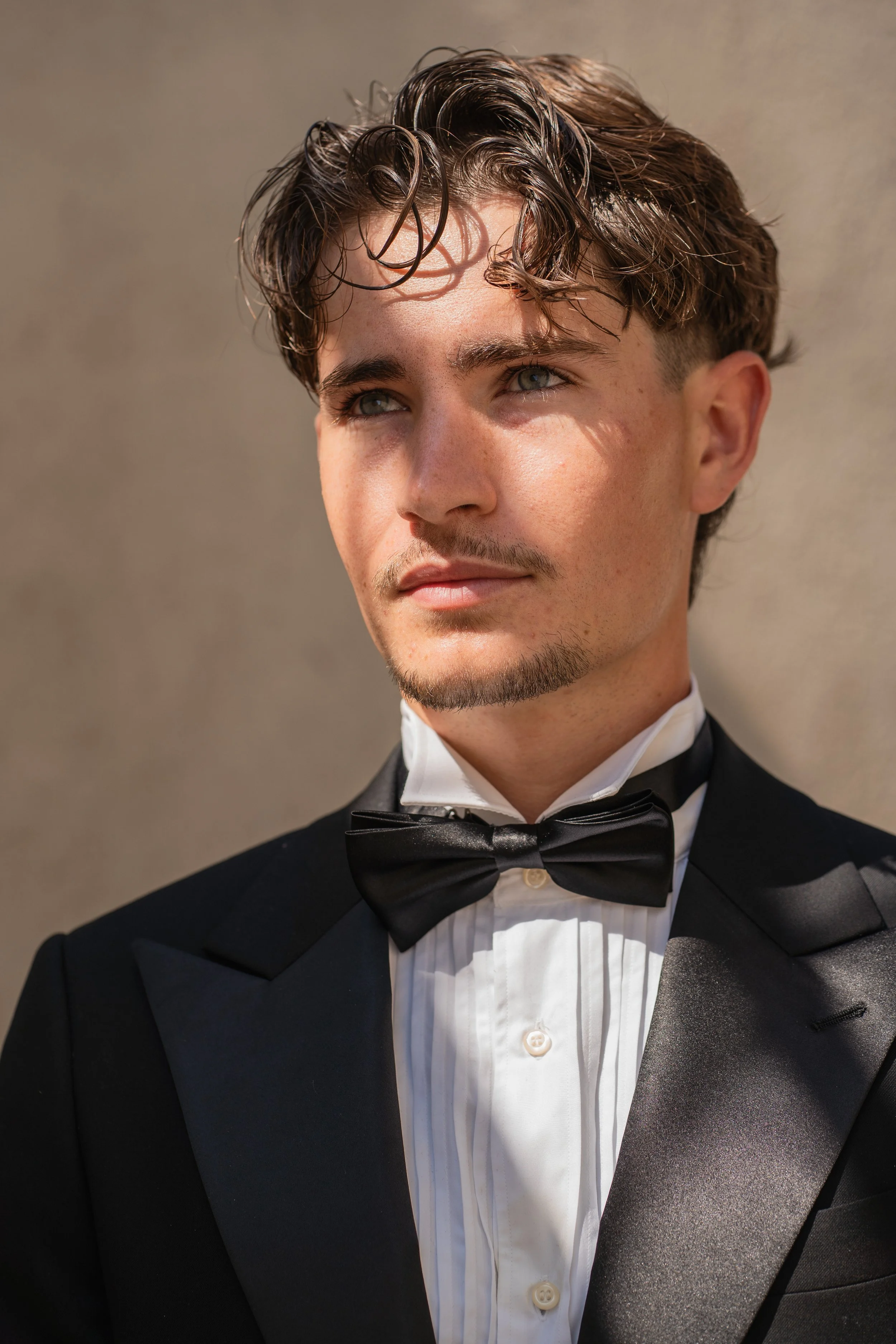 Close-up of a young man dressed in a black tuxedo with a white shirt and black bow tie, looking to his right with sunlight on his face.