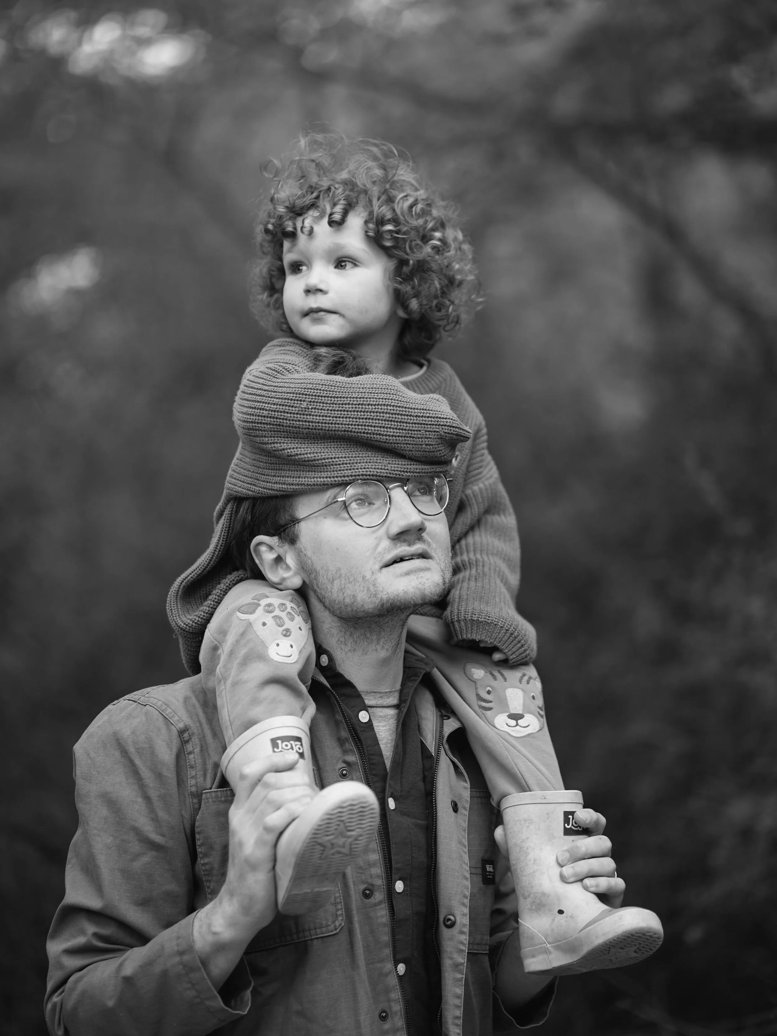 A man with glasses carrying a young curly-haired child on his shoulders, both holding coffee cups outdoors. The child looks to the side with a calm expression. The background is blurred with trees.