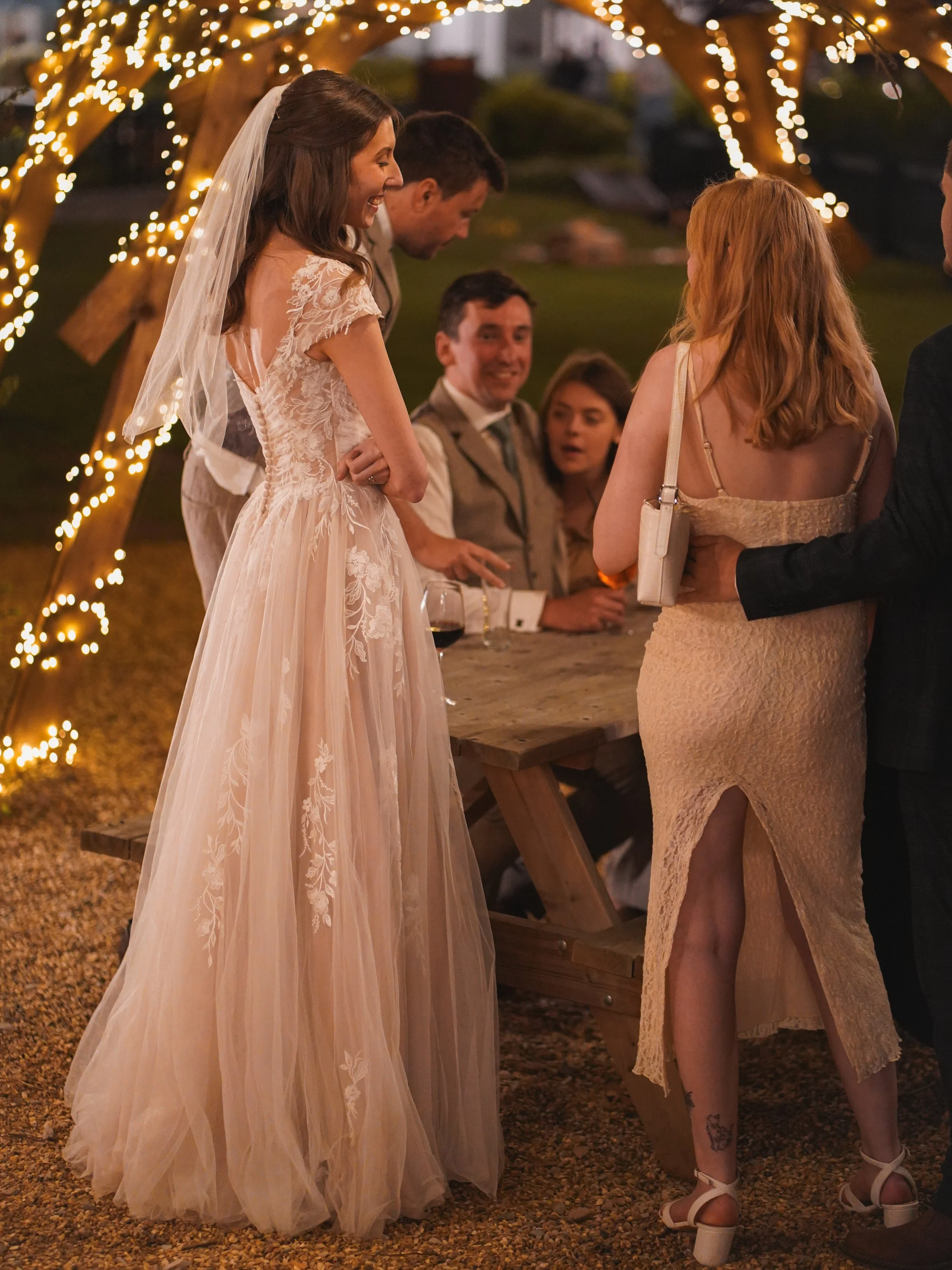 A bride in a lace wedding dress and veil smiling and talking to a red-haired woman in a beige dress at an outdoor wedding reception with string lights and wooden arch in the background.