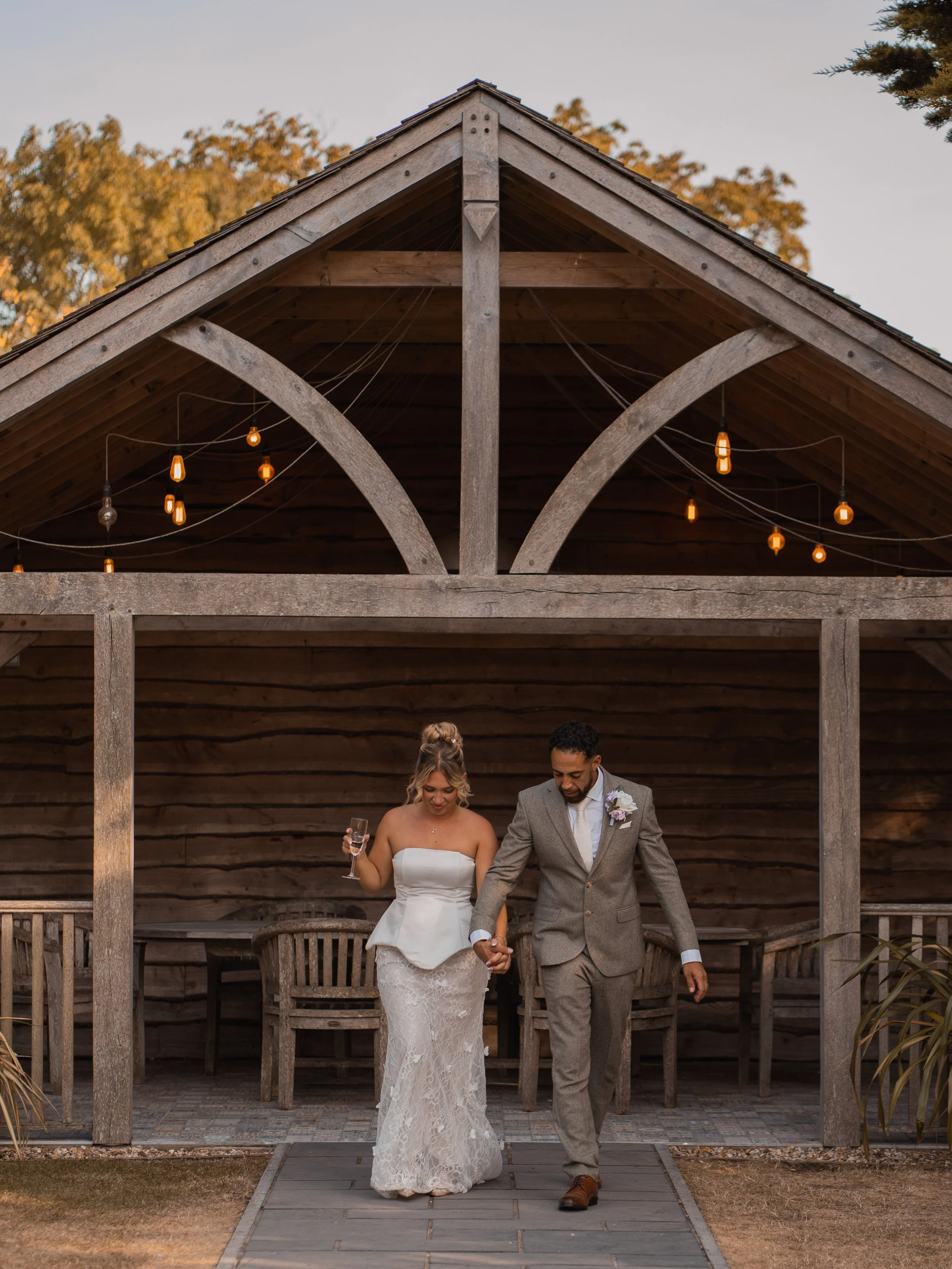 A bride and groom walk hand-in-hand outside a wooden structure, with the bride holding a champagne glass in her left hand. They are dressed in wedding attire, with trees and string lights hanging above them, in an outdoor setting during autumn.