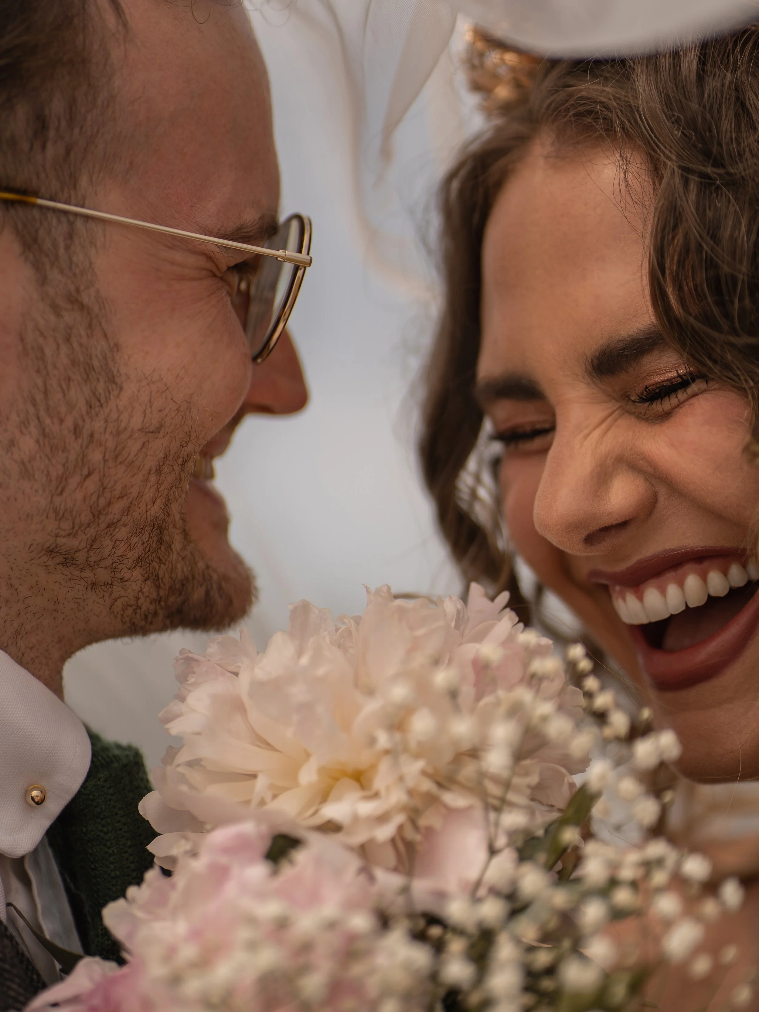 A close-up of a couple smiling and laughing with their foreheads touching, surrounded by a bouquet of white and pink flowers.
