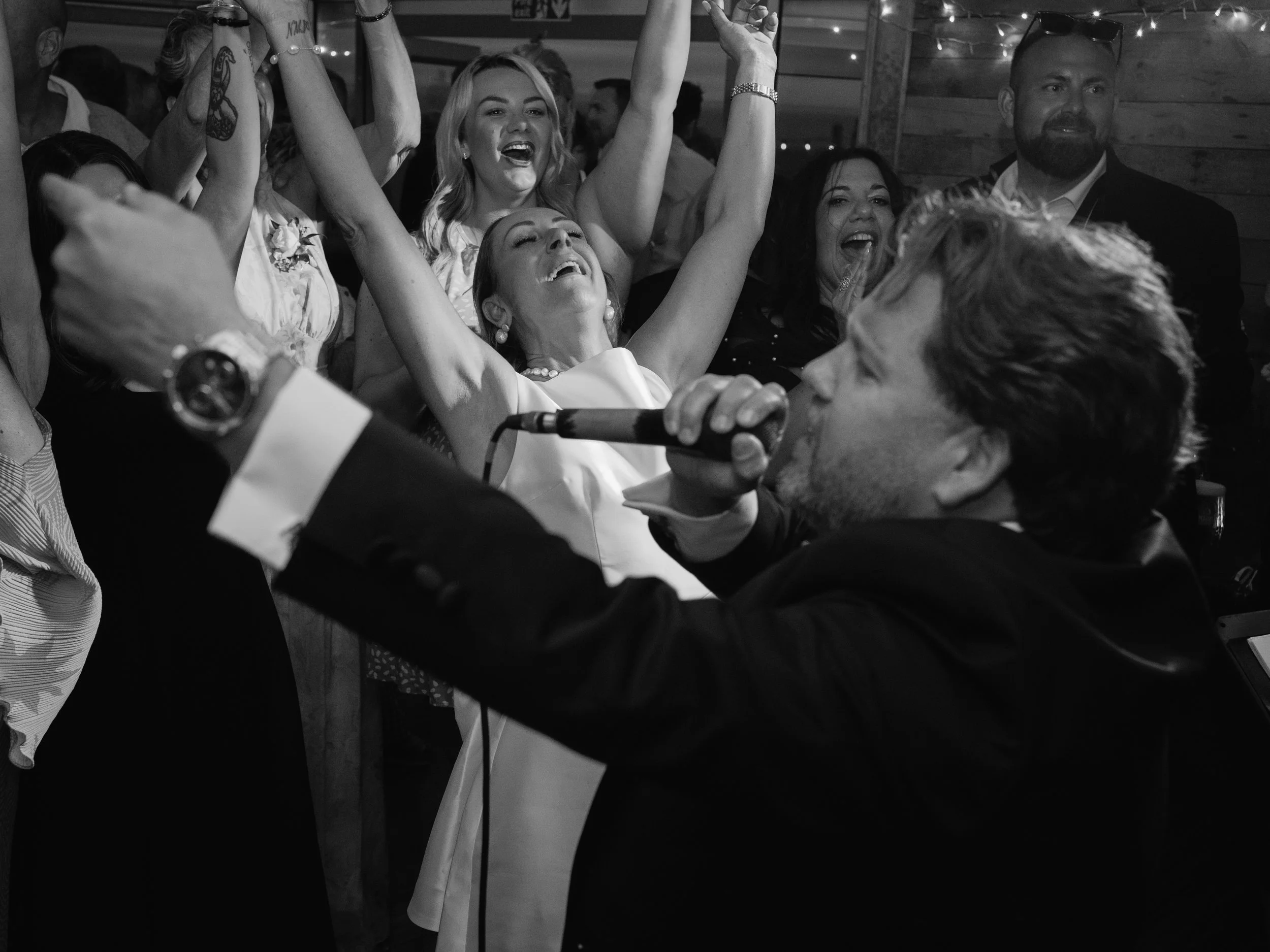 Group of people enjoying a lively celebration, some with raised arms and smiling faces, in a dimly lit venue with string lights, black and white photo.