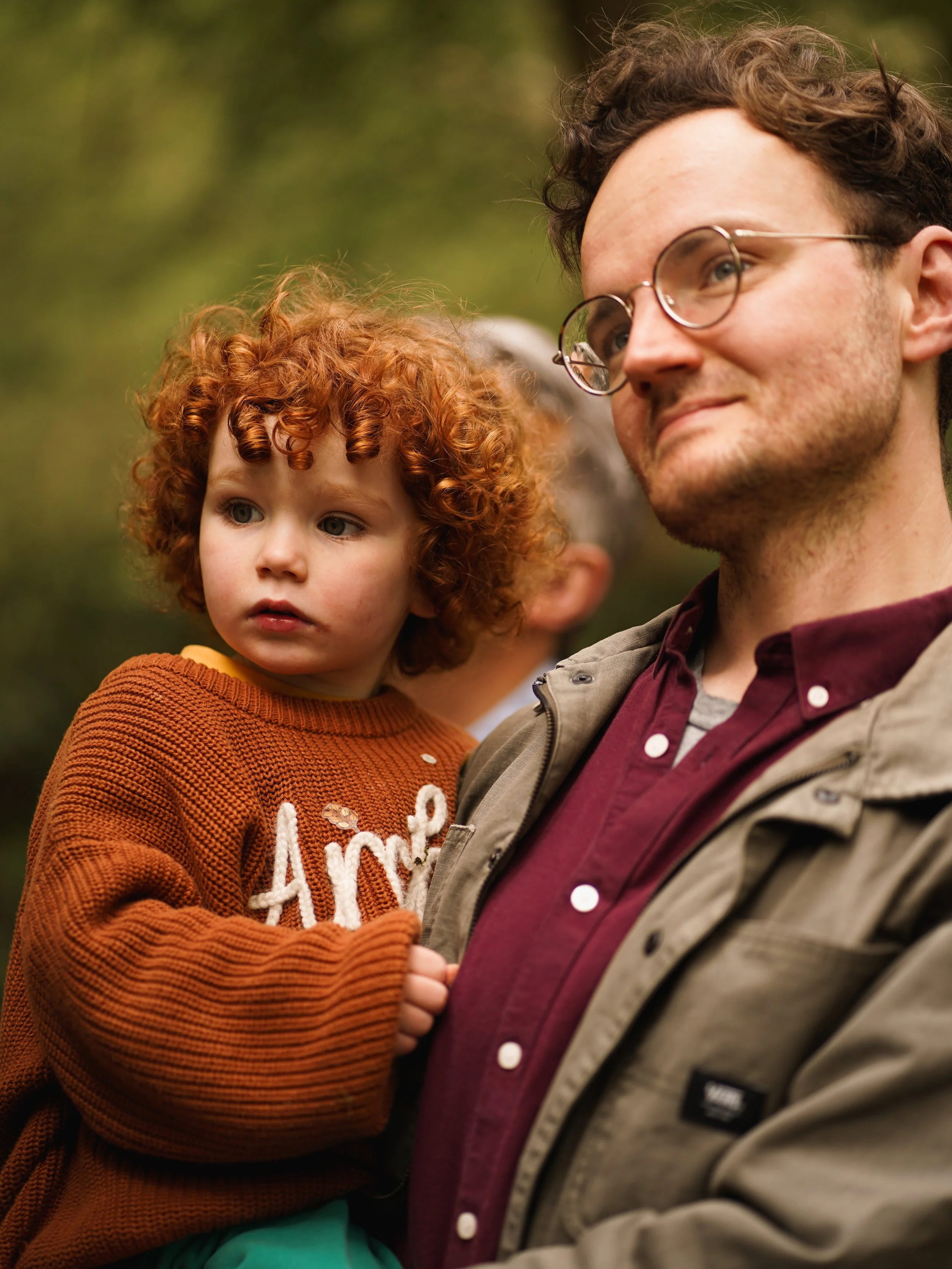 A man with glasses and curly hair standing outdoors next to a young girl with red curly hair. The girl is wearing an orange sweater and the man is dressed in a maroon shirt with a beige jacket.