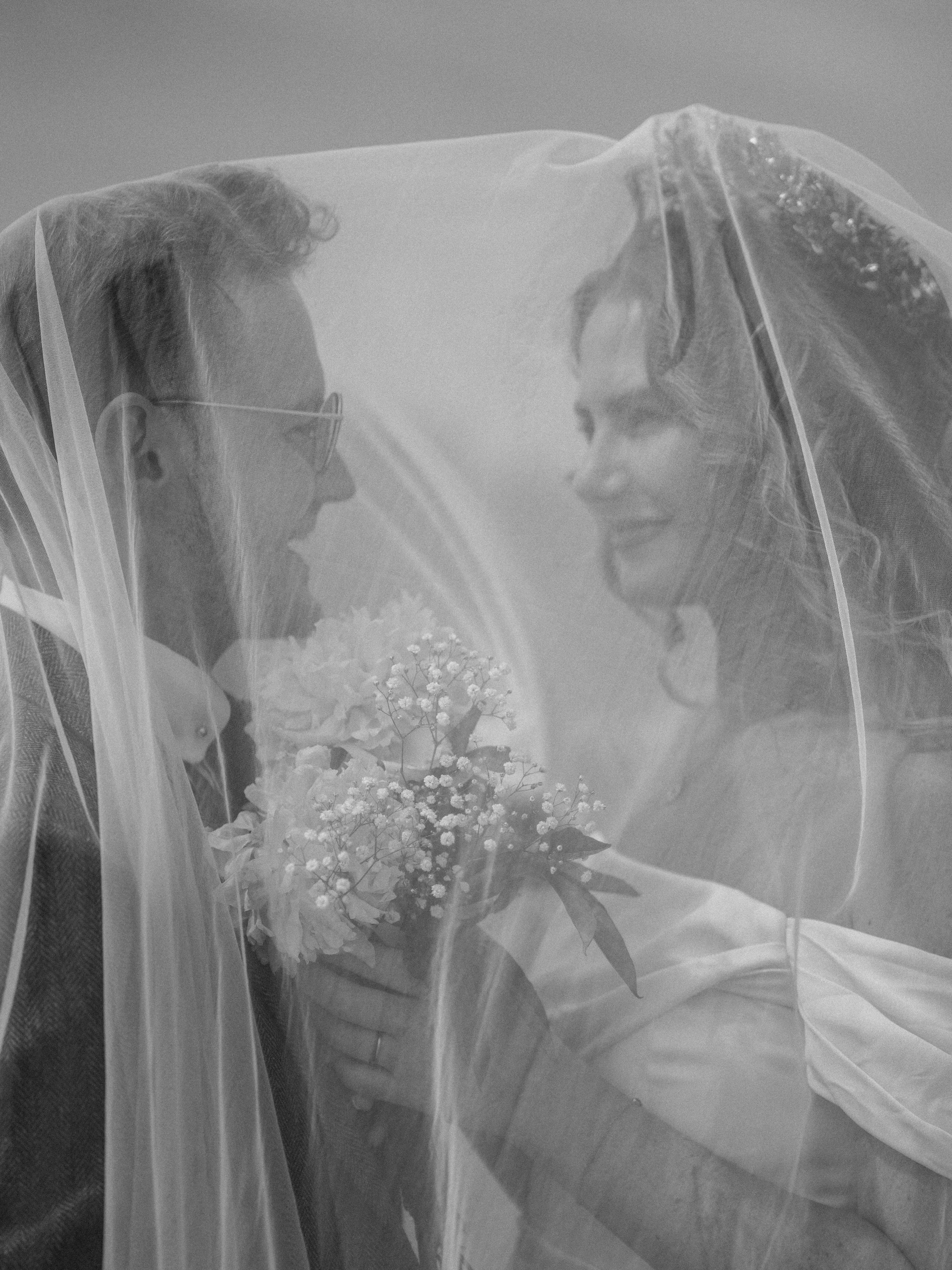 A bride and groom face each other under a veil, holding a bouquet of flowers.