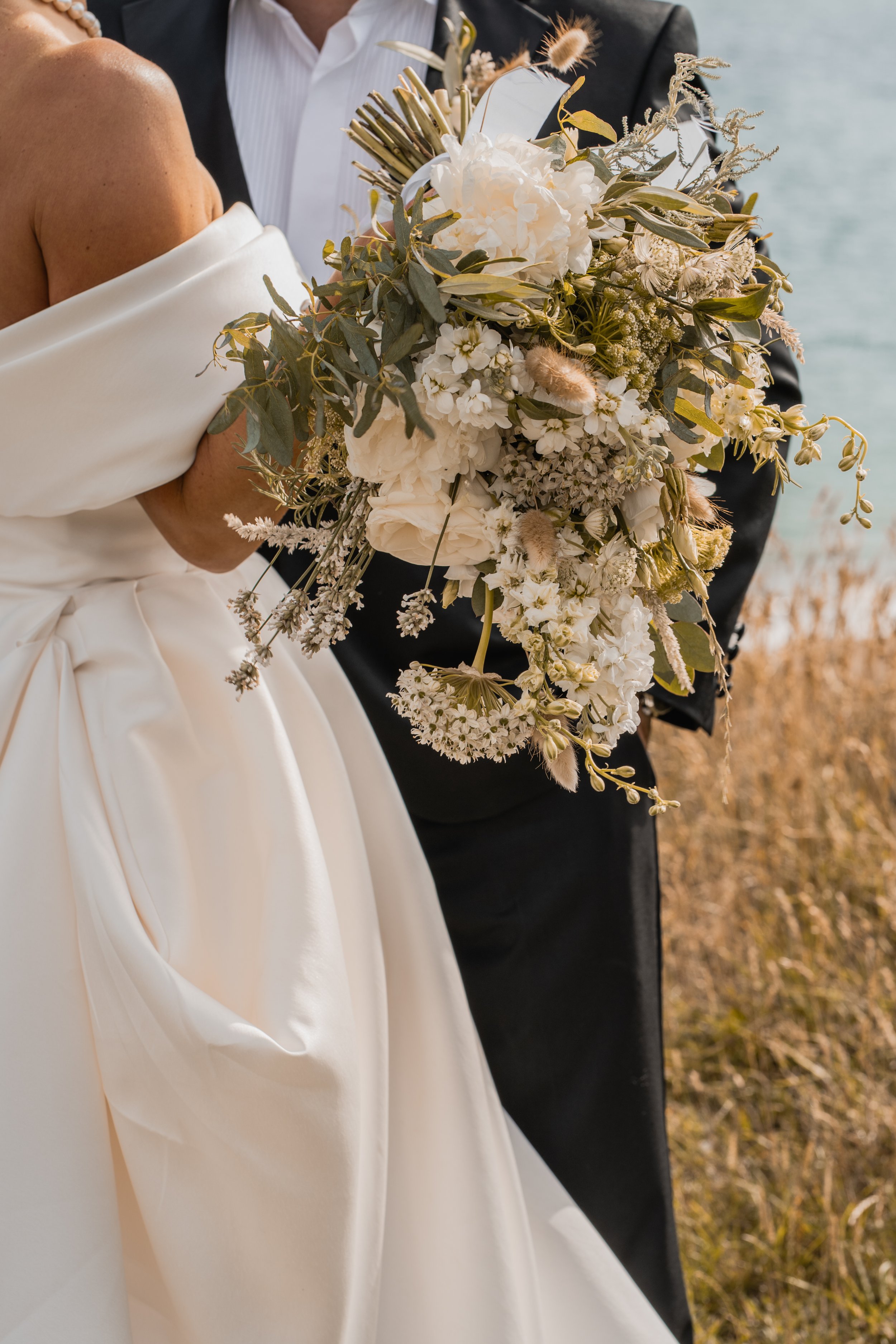 Close-up of a bride and groom holding a large cascading white and cream flower bouquet with greenery, against an outdoor natural background.