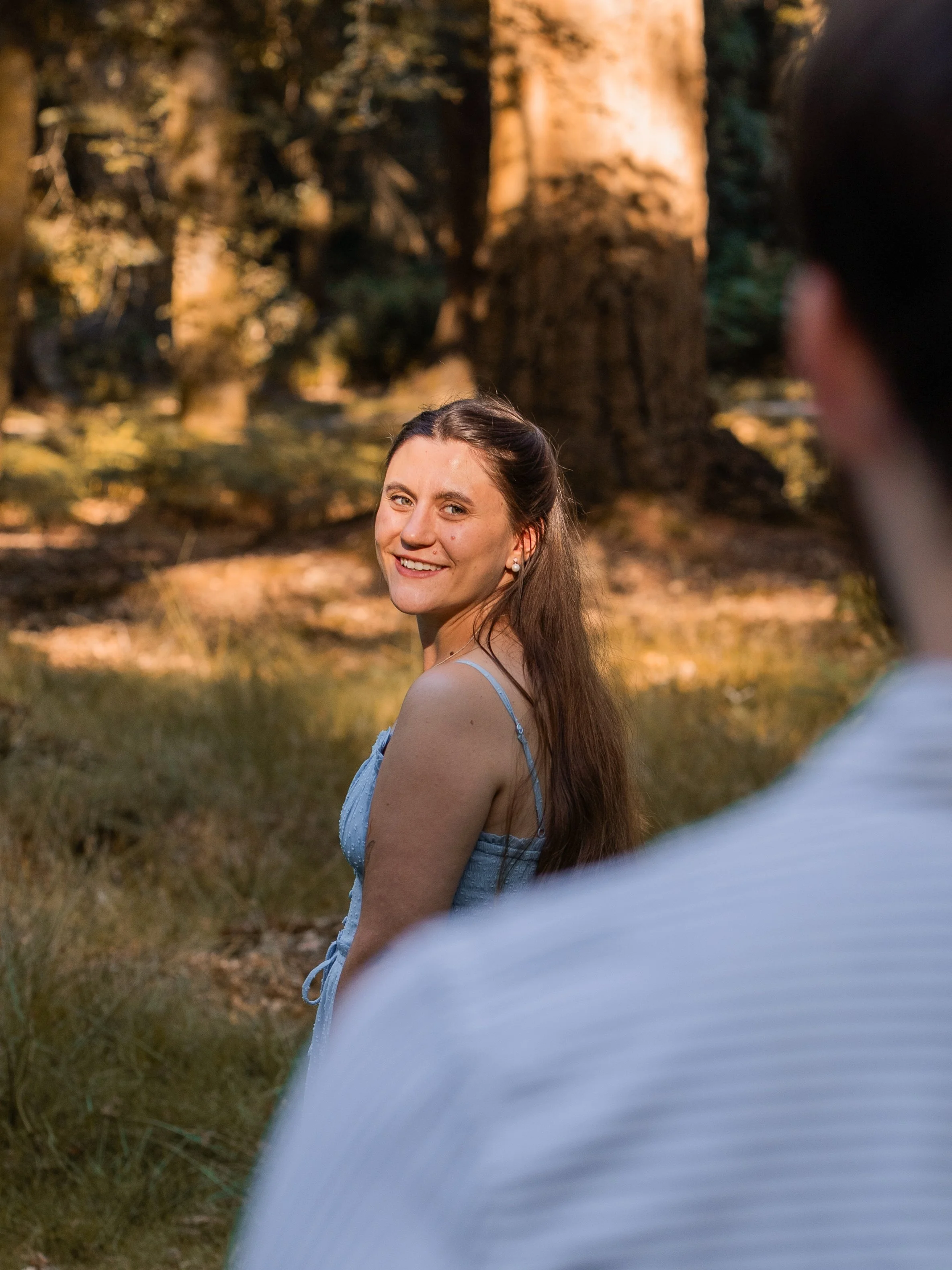 A woman with long brown hair wearing a blue dress smiling in a forested area during sunset, with part of another person's shoulder and arm visible in the foreground.