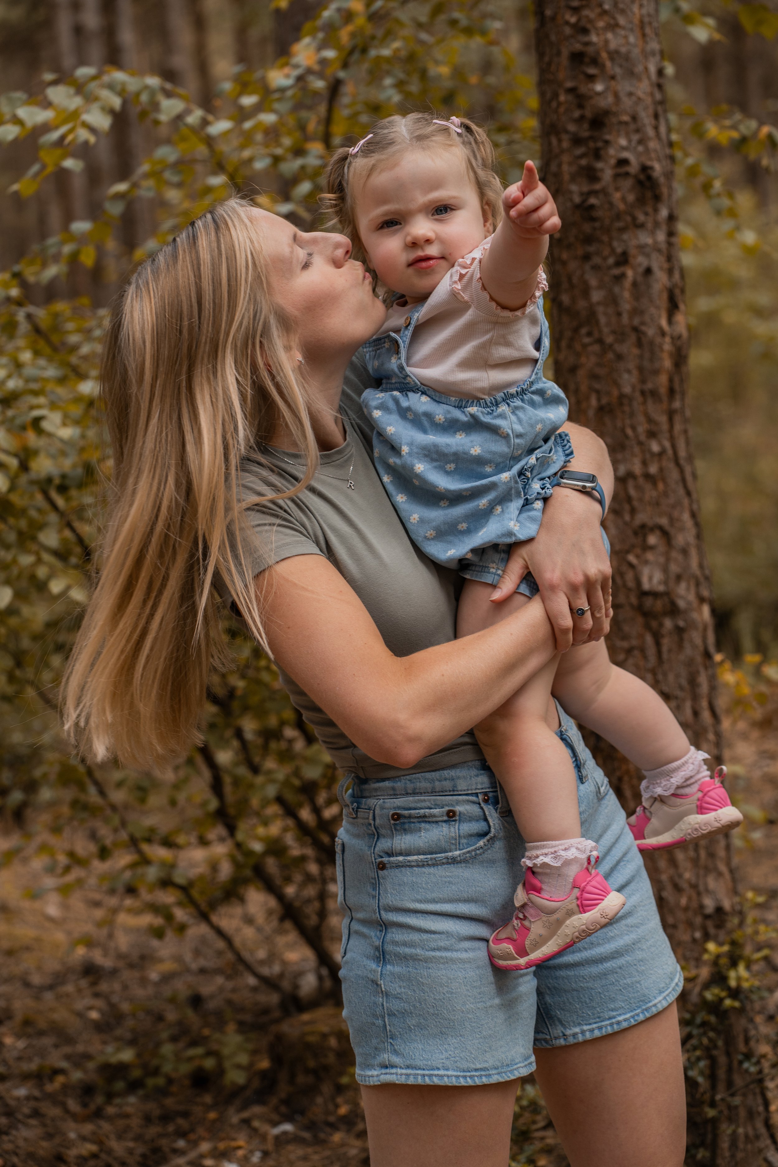 A woman holding a young girl in a forest with autumn leaves, the girl pointing at something in the distance.