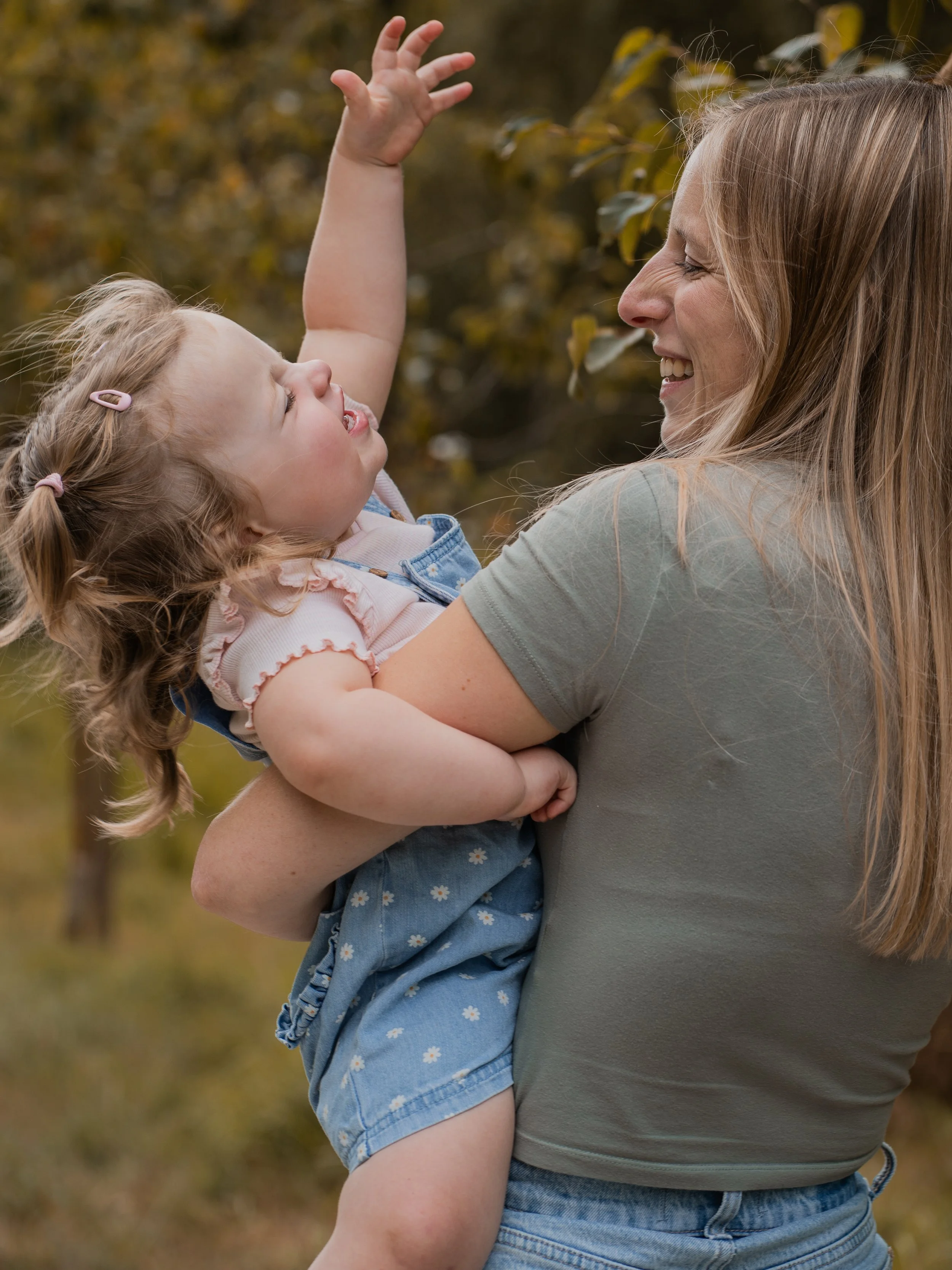 A woman smiling and holding a young girl outdoors during fall, with autumn leaves in the background.