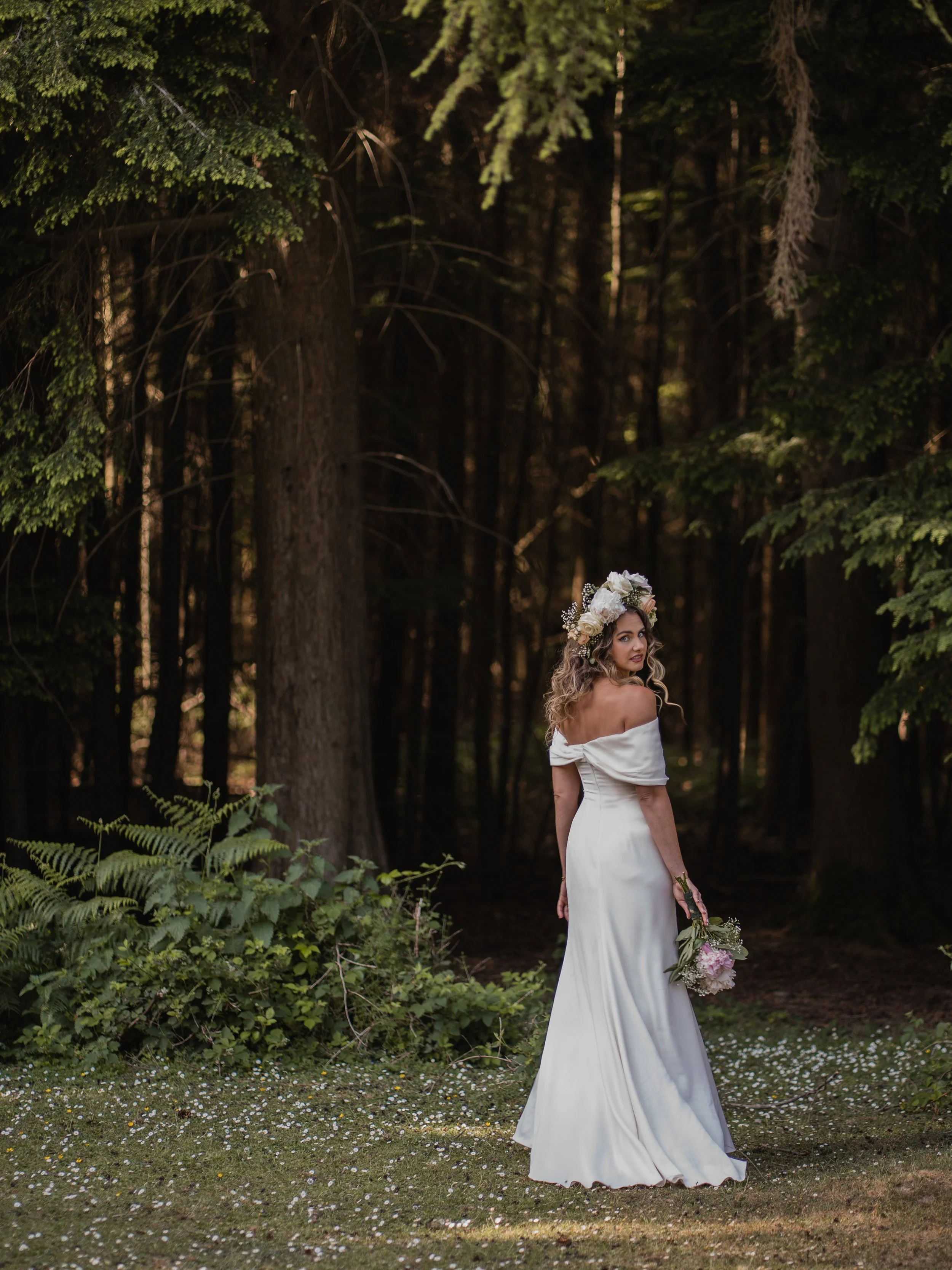 A woman in a white off-shoulder dress holding a bouquet of flowers standing in a forest.