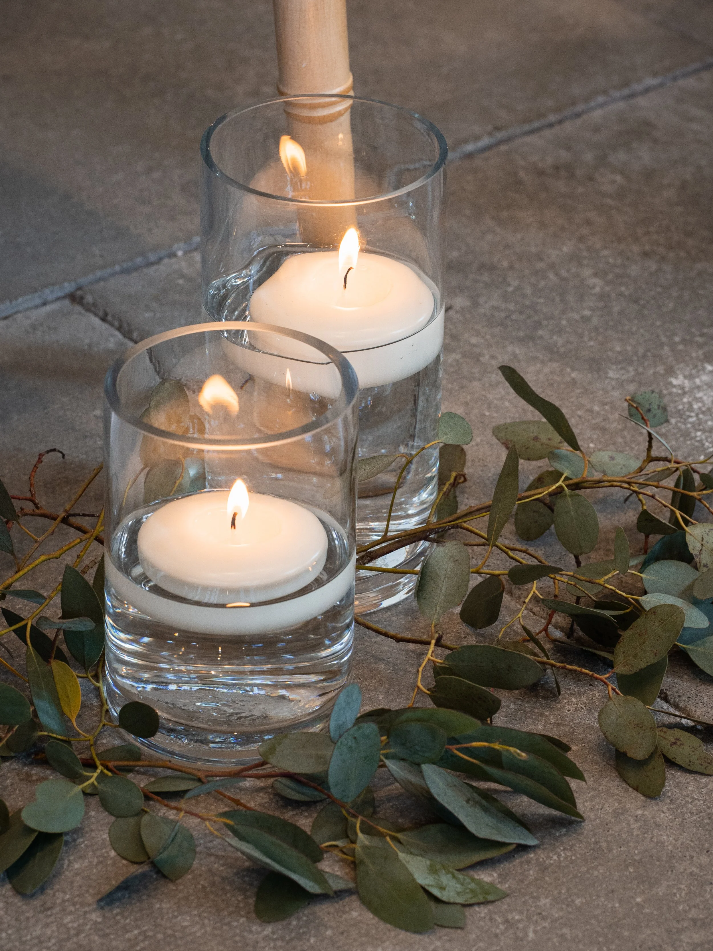 Three glass candle holders with lit white candles on a concrete surface, surrounded by eucalyptus leaves.