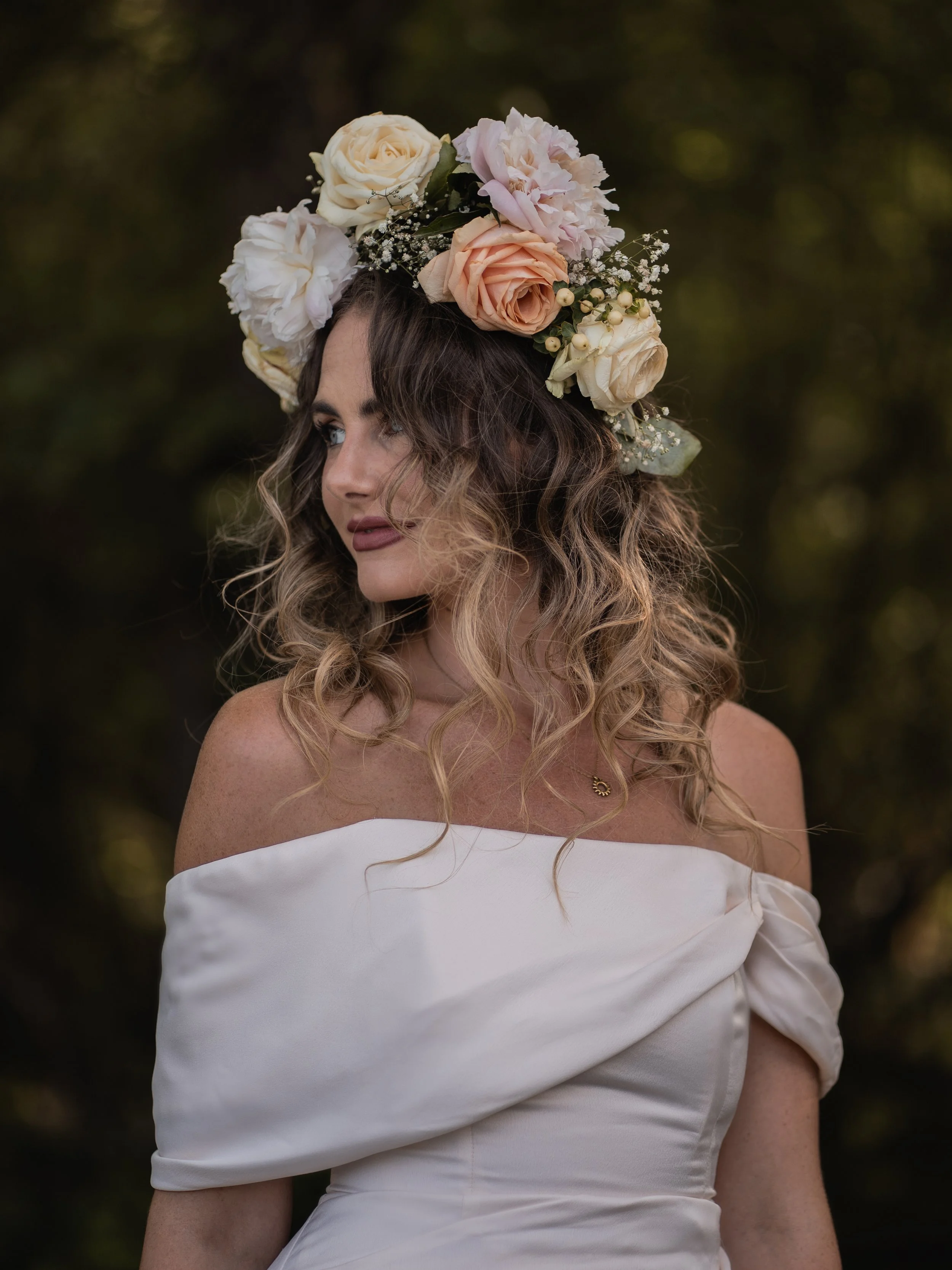 A woman with long, wavy hair wearing an off-shoulder white dress and a large floral crown made of white, peach, and pink roses, along with small white filler flowers, standing outdoors with a dark, blurry background.
