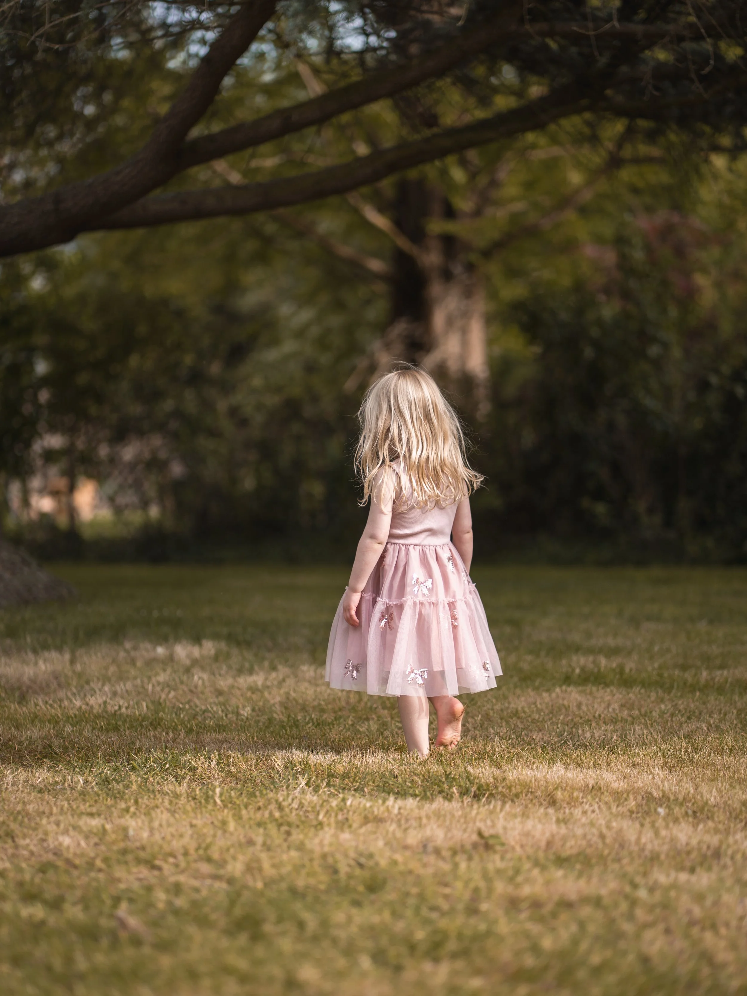 A young girl with long blonde hair wearing a pink dress walking barefoot on grass in a park with tall trees and lush greenery. Editorial family photoshoot. 