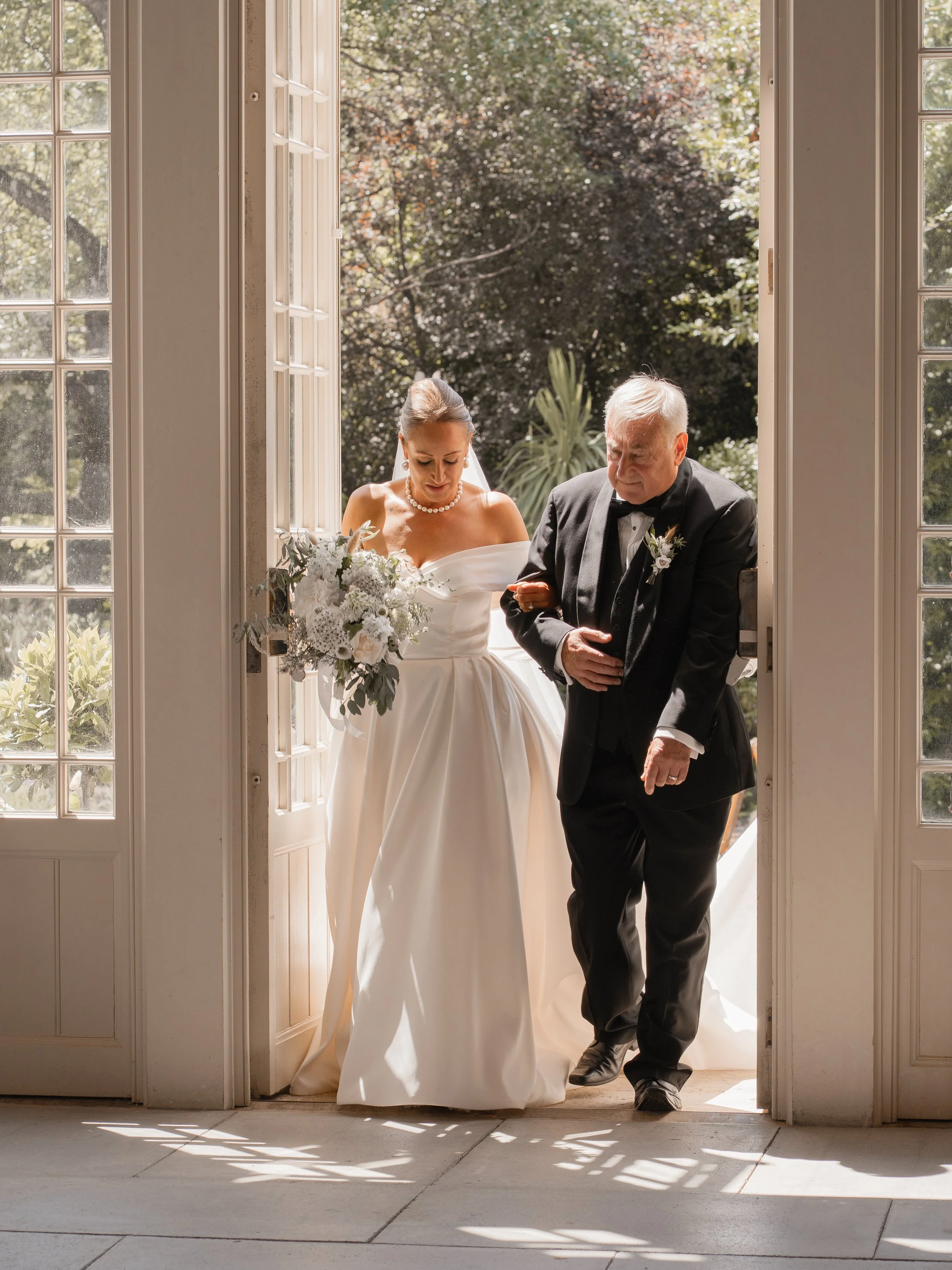 A bride in a white wedding dress holding a bouquet of flowers, walking out of a door with an older man in a suit. The background shows trees and sunlight.
