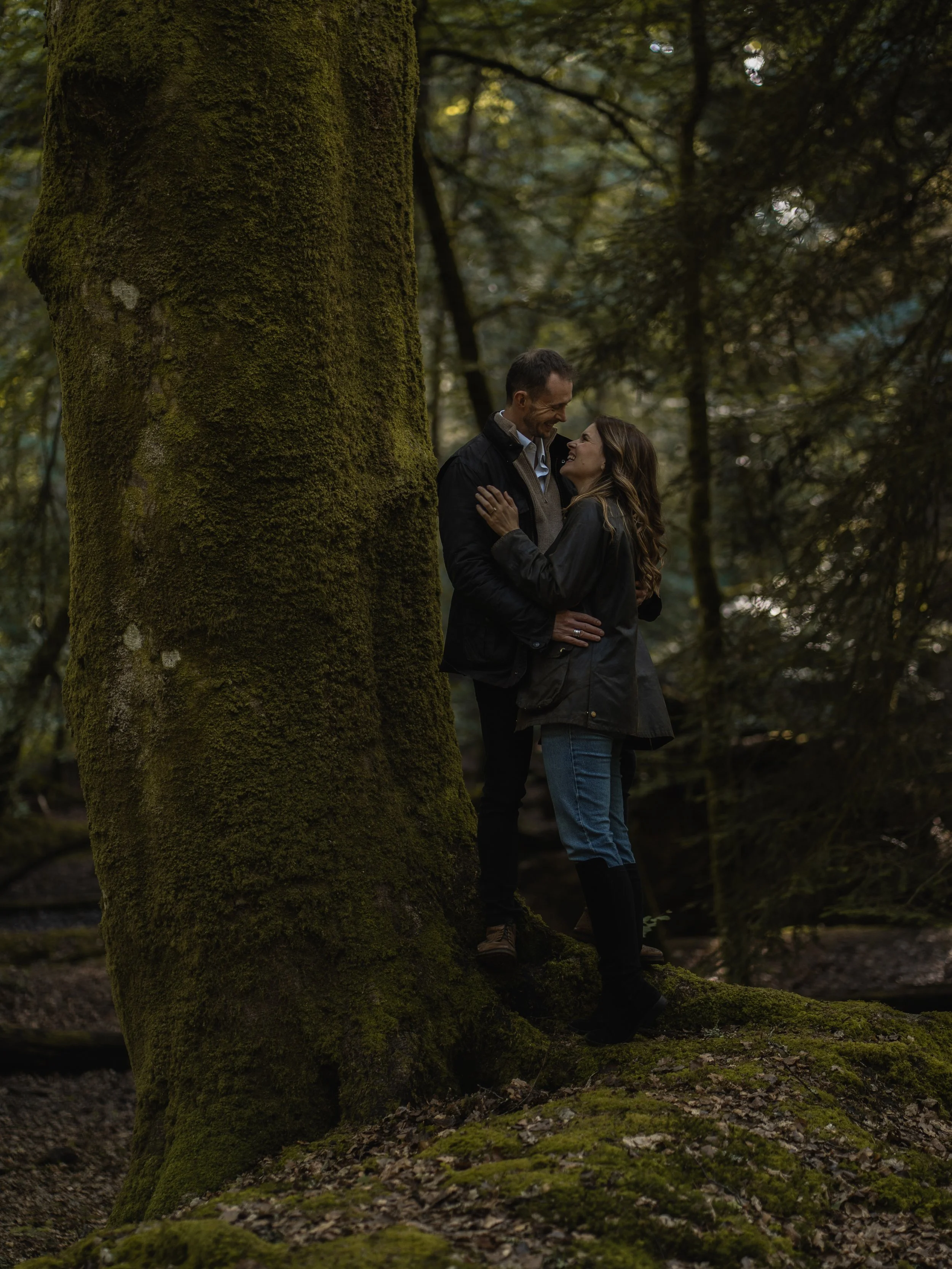 A couple standing close together and smiling at each other in a forest with large moss-covered trees and a forest floor covered with moss and leaves.