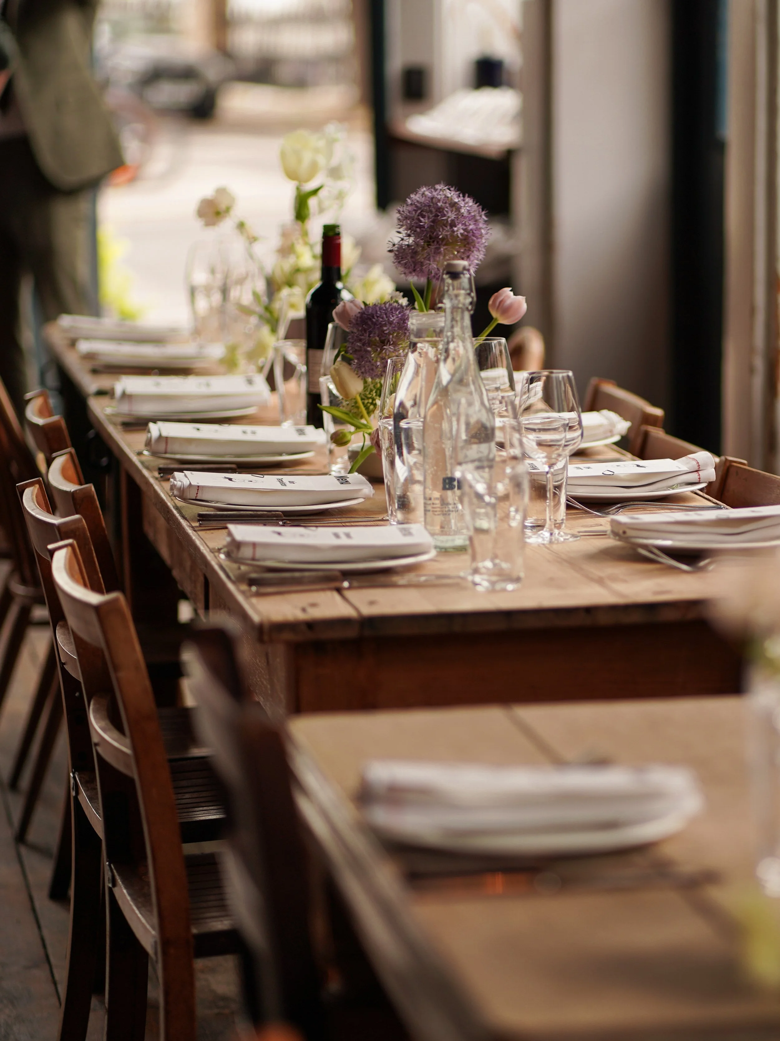 A long wooden table set for a meal with wine bottles, water bottles, glasses, and floral centerpieces, situated indoors near a window with a view of the outdoors.