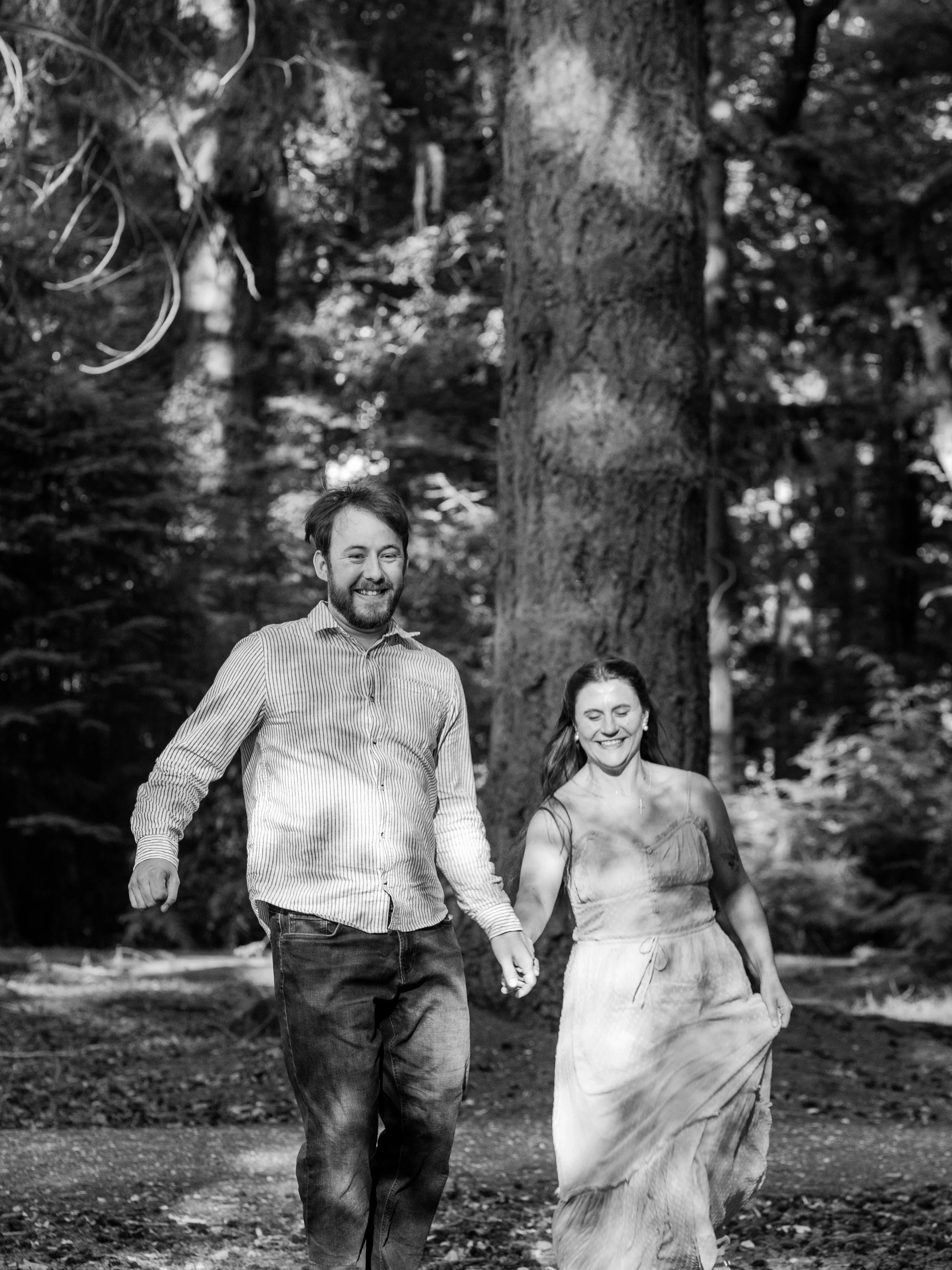 A man and a woman holding hands and walking together in a forest. The man has a beard, is smiling, and wearing a striped shirt and jeans. The woman has long hair, is also smiling, and wearing a long skirt and top. Tall trees surround them, with sunli