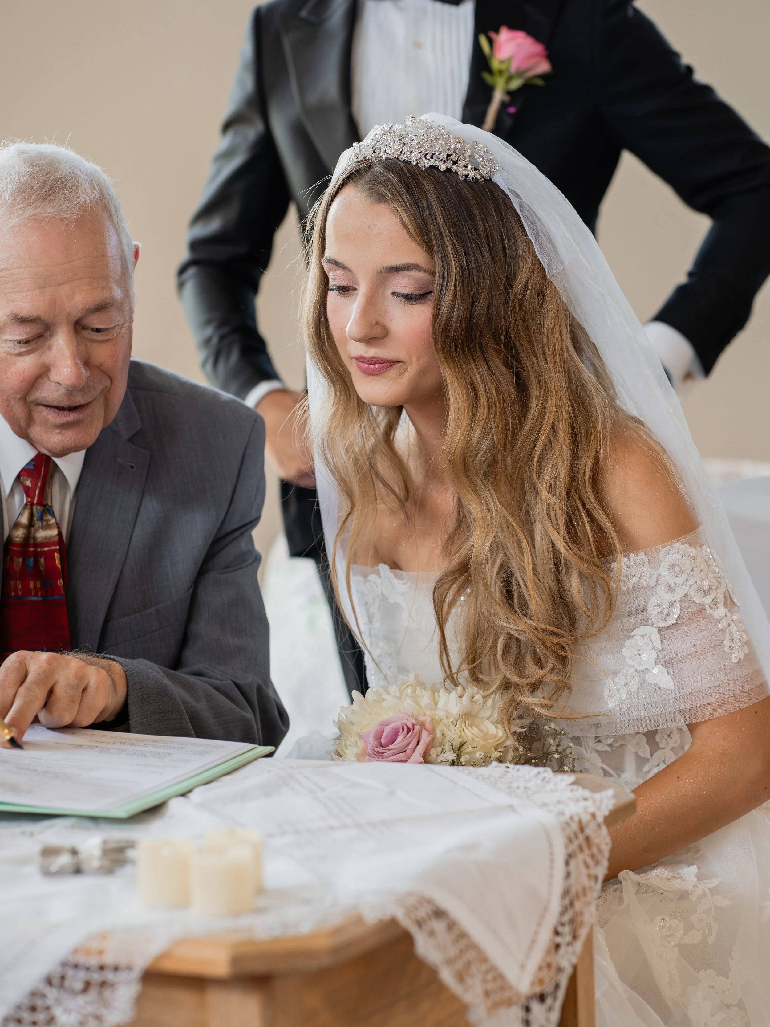 A bride with long wavy hair, wearing a tiara and veil, in a wedding dress with lace details, sitting at a table with an older man in a suit, signing a document. A person in a black suit with a pink flower boutonniere stands behind them. The table is 