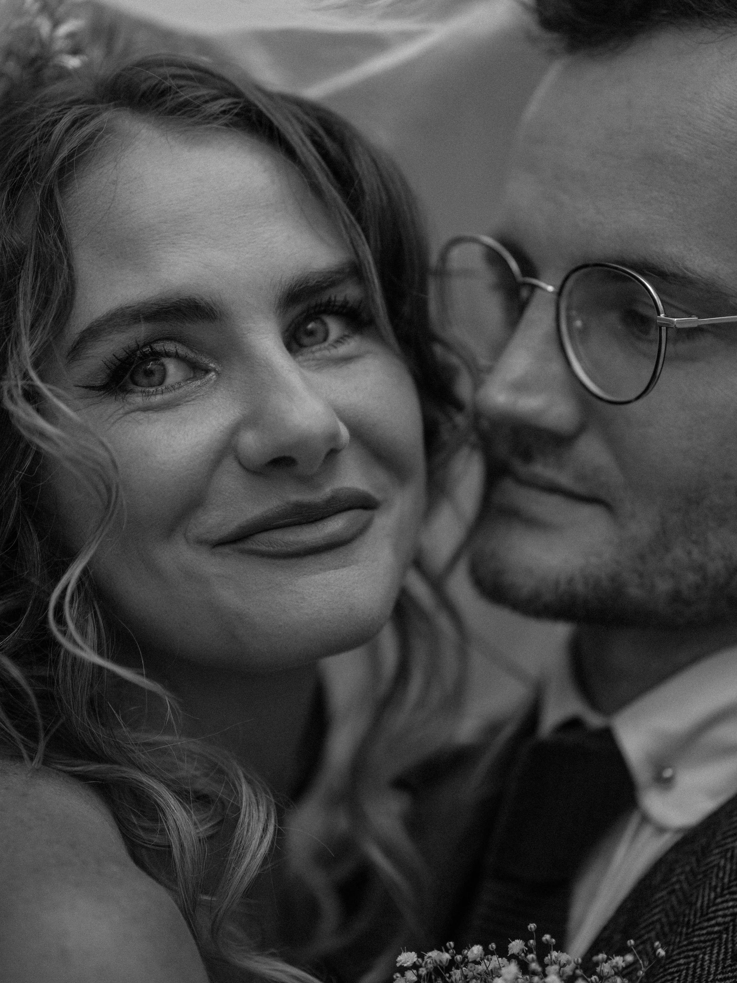 A black and white close-up photo of a woman with wavy hair and bright eyes, looking at a man with glasses who is holding a bouquet of small flowers.
