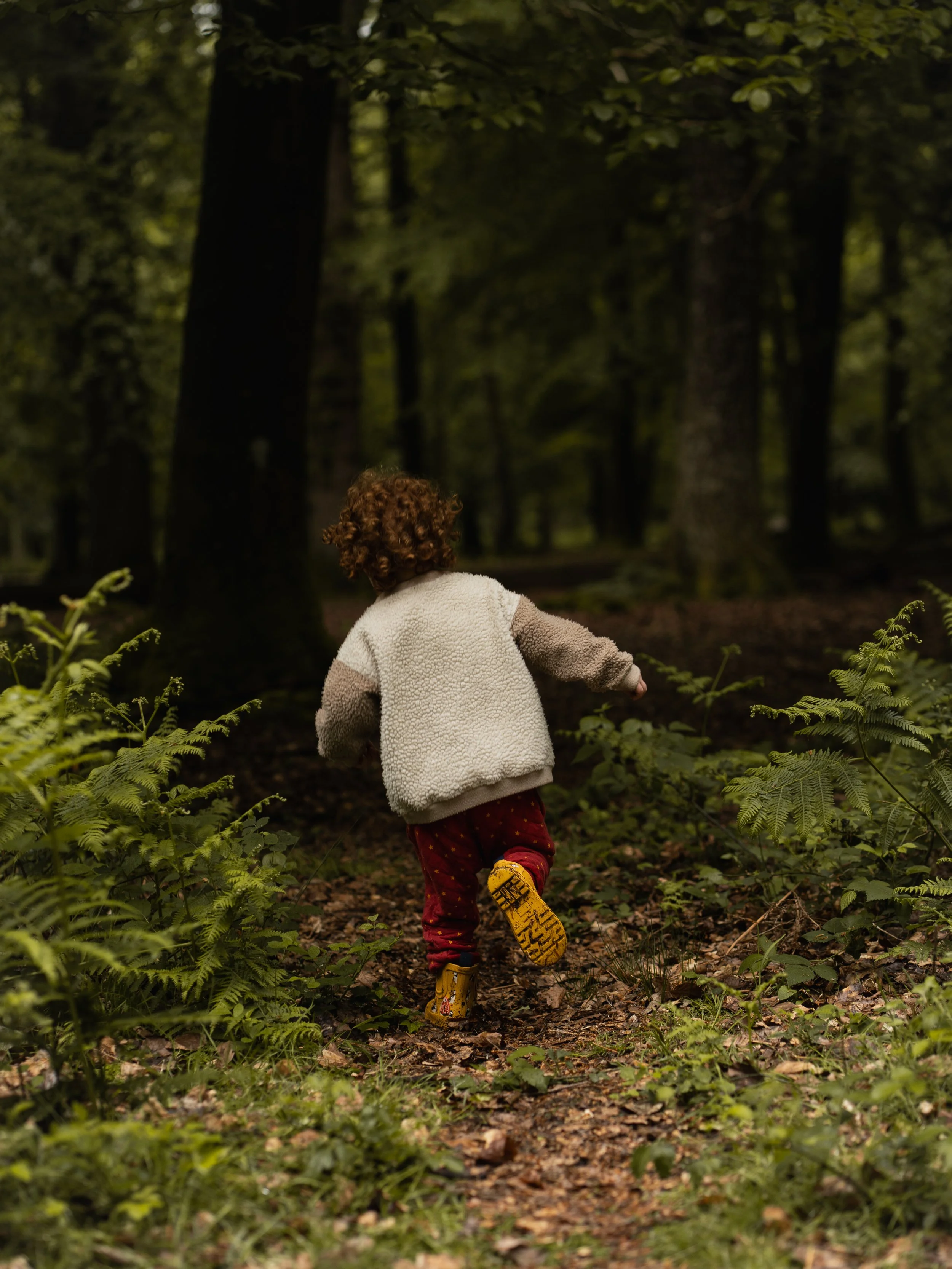 Child wearing a beige and brown fleece jacket, red pants with yellow patterns, and yellow boots, running through a dense forest trail surrounded by green foliage and tall trees.