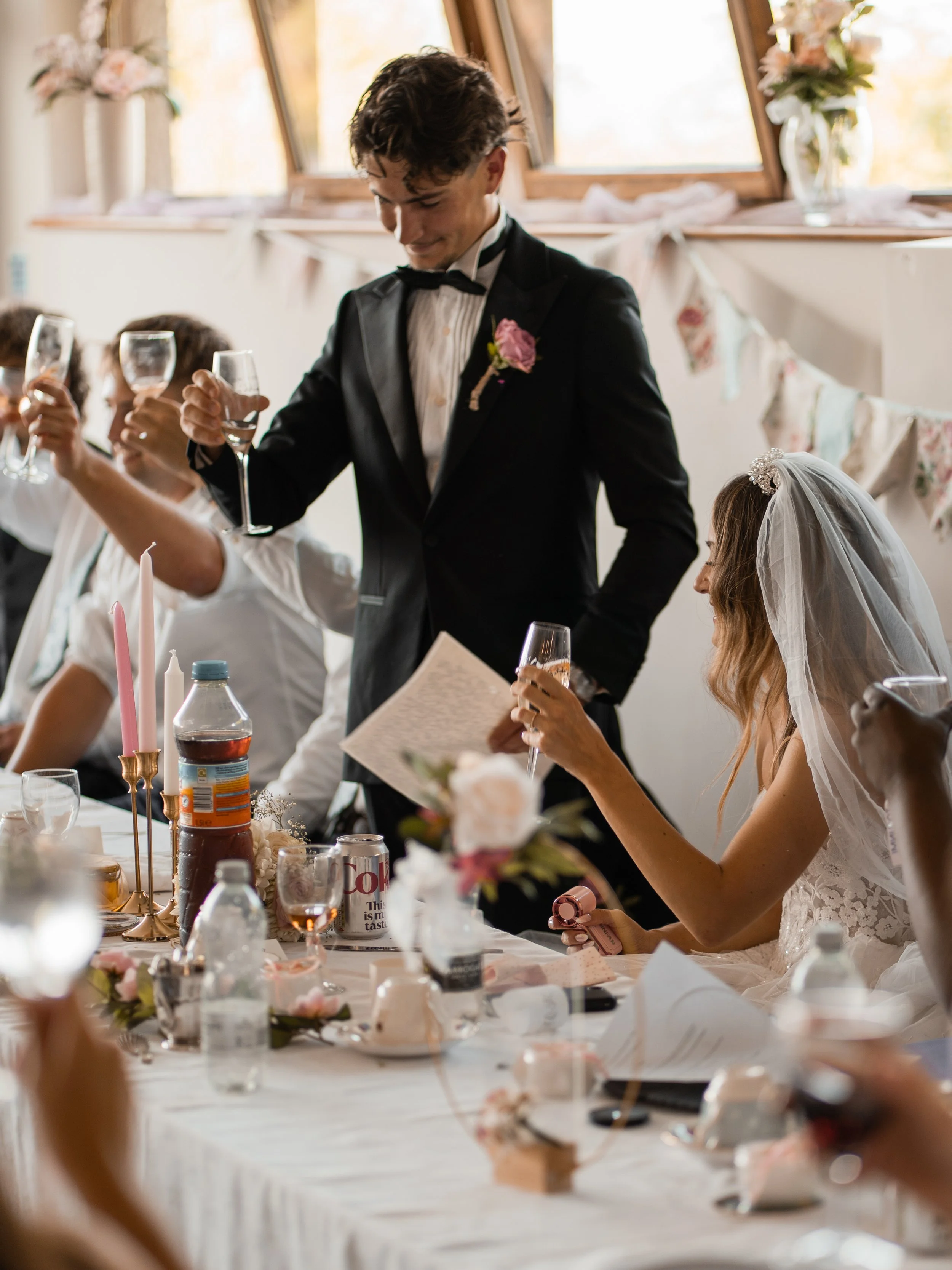A groom in a tuxedo holding a glass of champagne at a wedding reception, with a woman in a wedding dress sitting at the table, surrounded by decorations and guests.