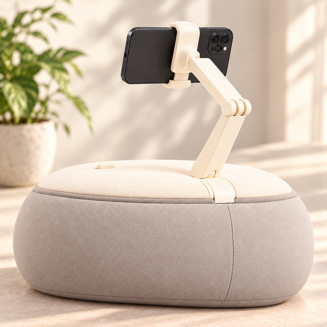 A smartphone mounted on a beige adjustable stand placed on a soft, rounded ottoman in a bright room with natural light and a potted plant in the background.