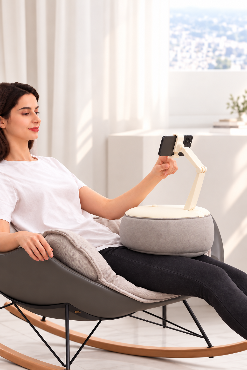 Woman seated in a modern rocking chair, holding a tablet mounted on a robotic arm, in a bright room with white curtains and a cityscape view.