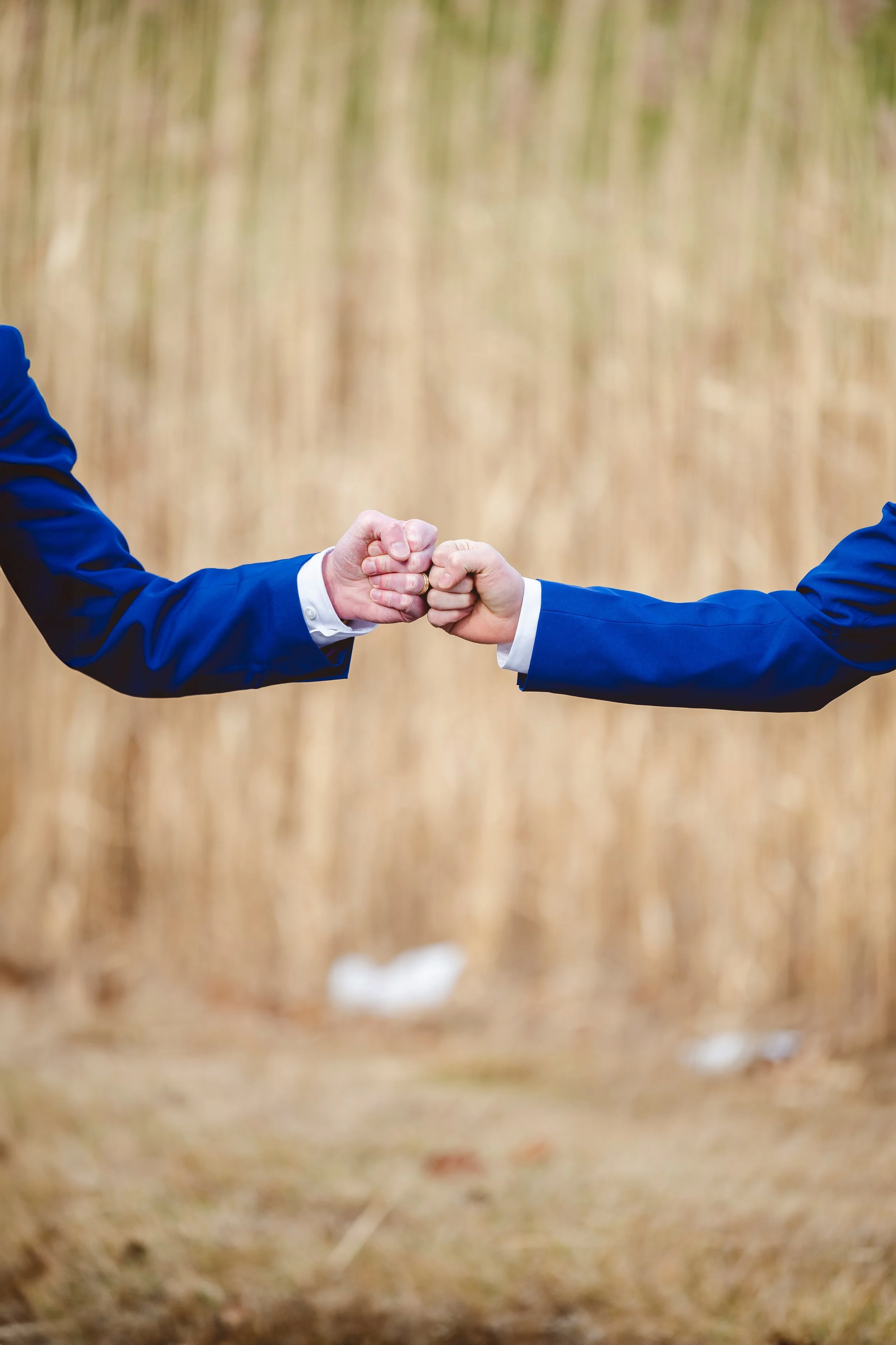 Two people in suits giving each other a fist bump in a natural outdoor setting.