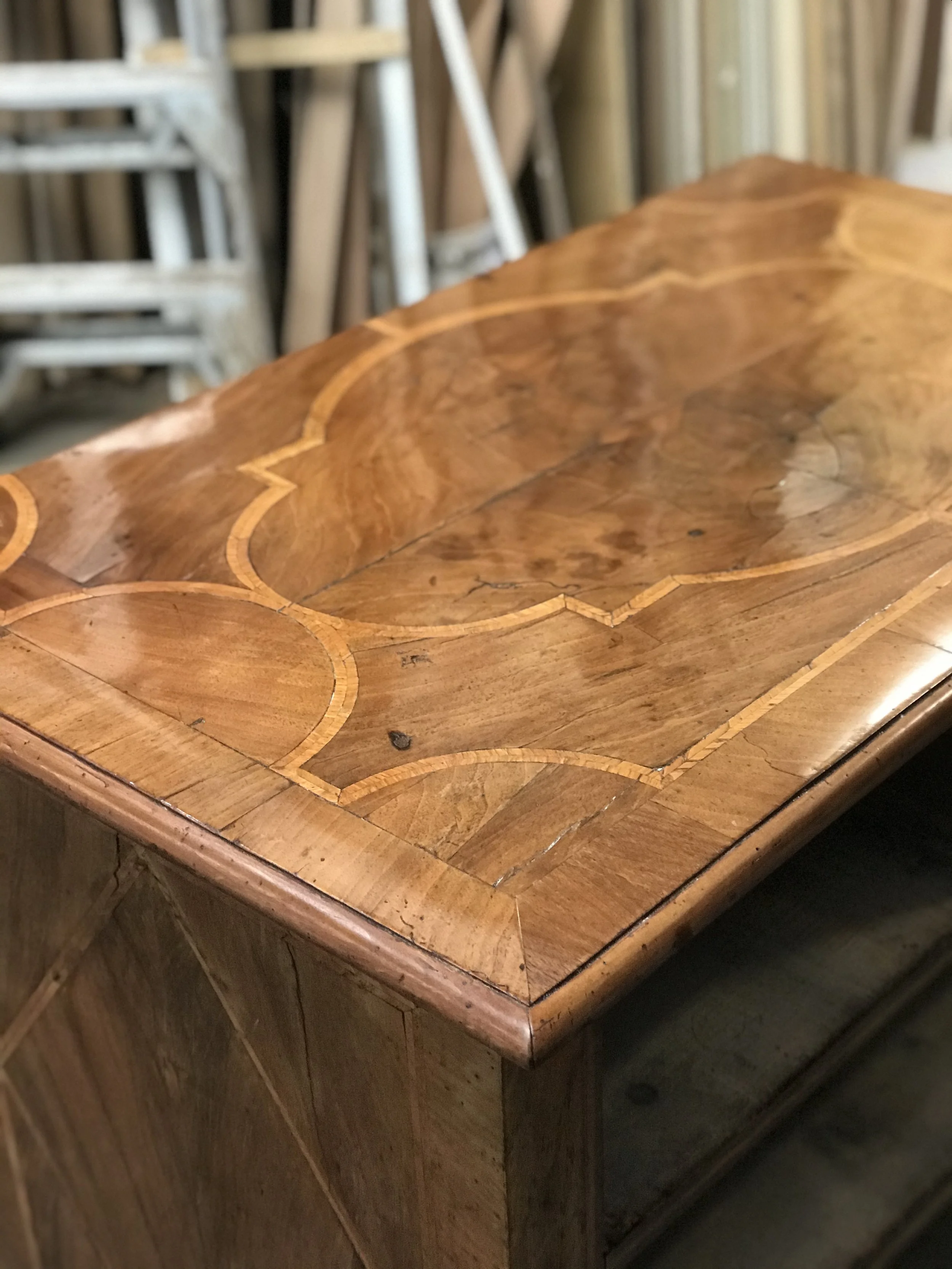 Close-up of a wooden table with intricate inlays and a polished surface, with a background of stacked wood and furniture in a workshop.
