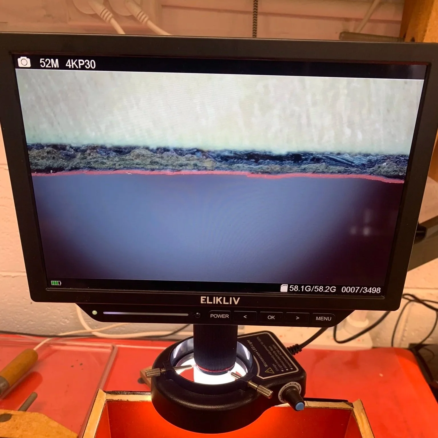 A digital microstop monitor displaying a cross section of lacquer on a box. The monitor has a black border and stands on a flexible arm on an orange work surface with various tools and wires nearby.