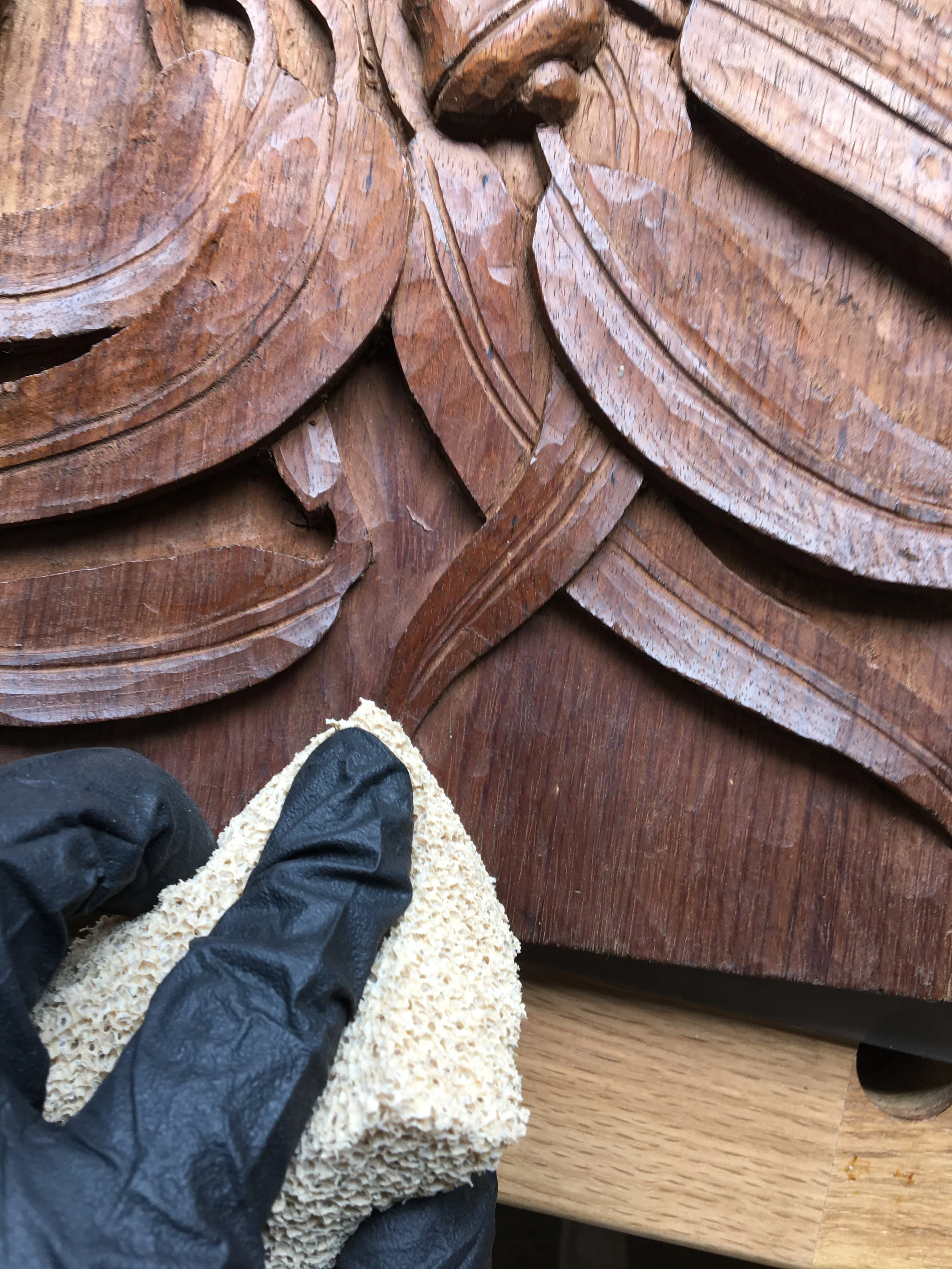 Close-up of a woodworking project showing detailed carved wooden designs, with a person's feet visible standing on a textured beige mat. The person is wearing black jacket and black shoes, standing near a polished wooden surface.