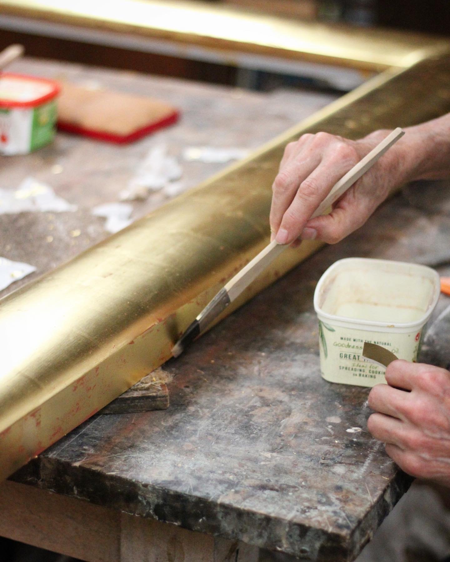 Hand holding a butter knife and spreading butter from a plastic butter container on a golden metal surface, with a table in the background.