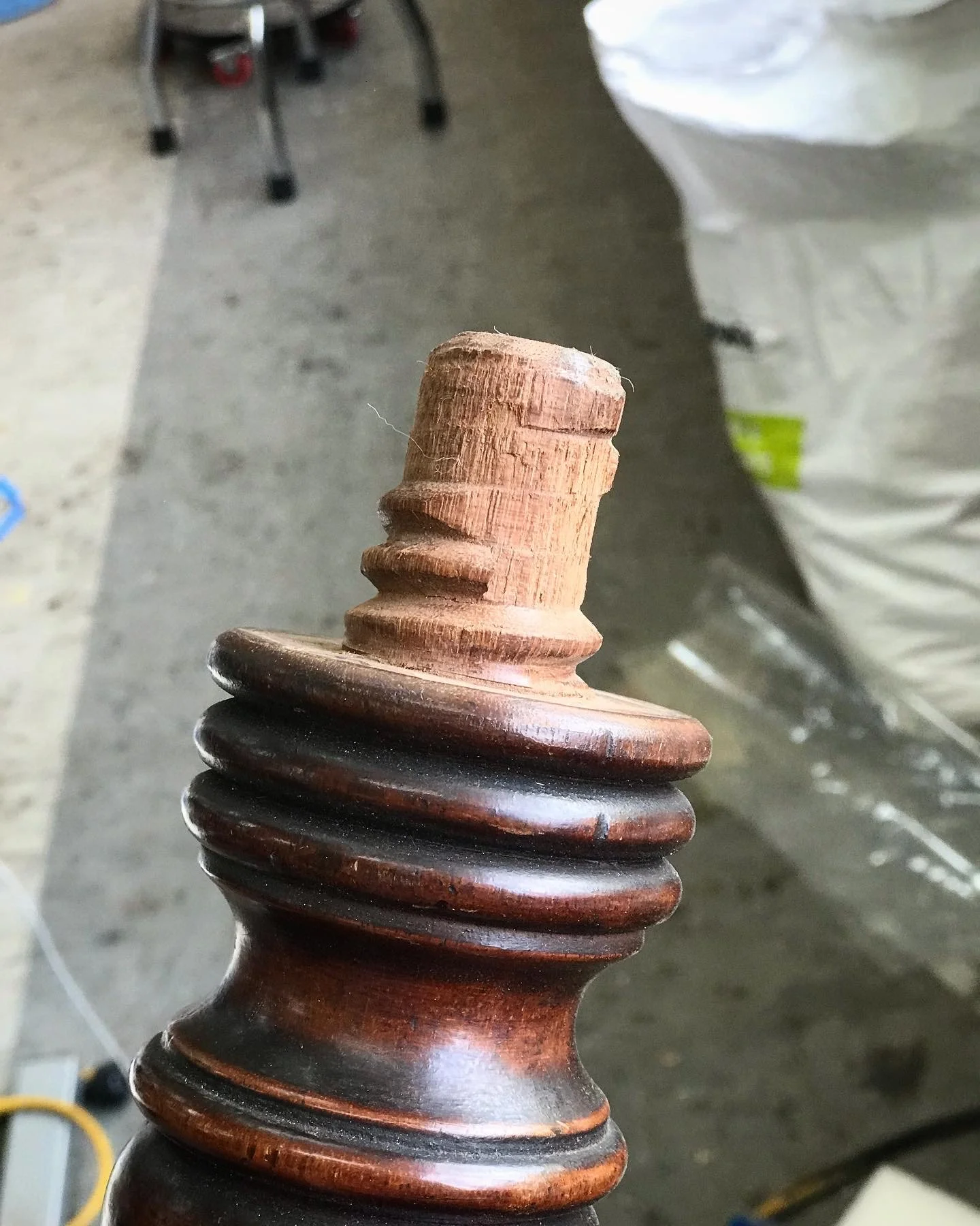 Close-up of a wooden chair leg with a heavily damaged wooden screw fitting, showing a background of a concrete floor and various objects.