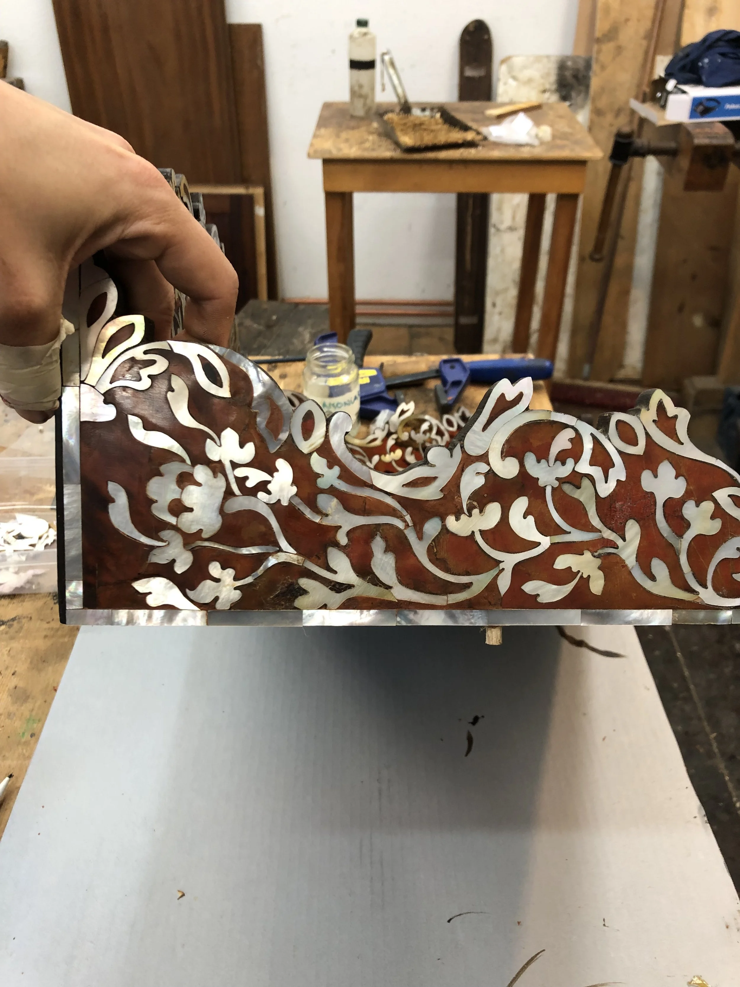 A person is holding a decorative, wood piece with floral patterns cut from turtle shell and abalone in front of a workbench in a woodworking or craft shop. The workbench has various tools and supplies in the background.