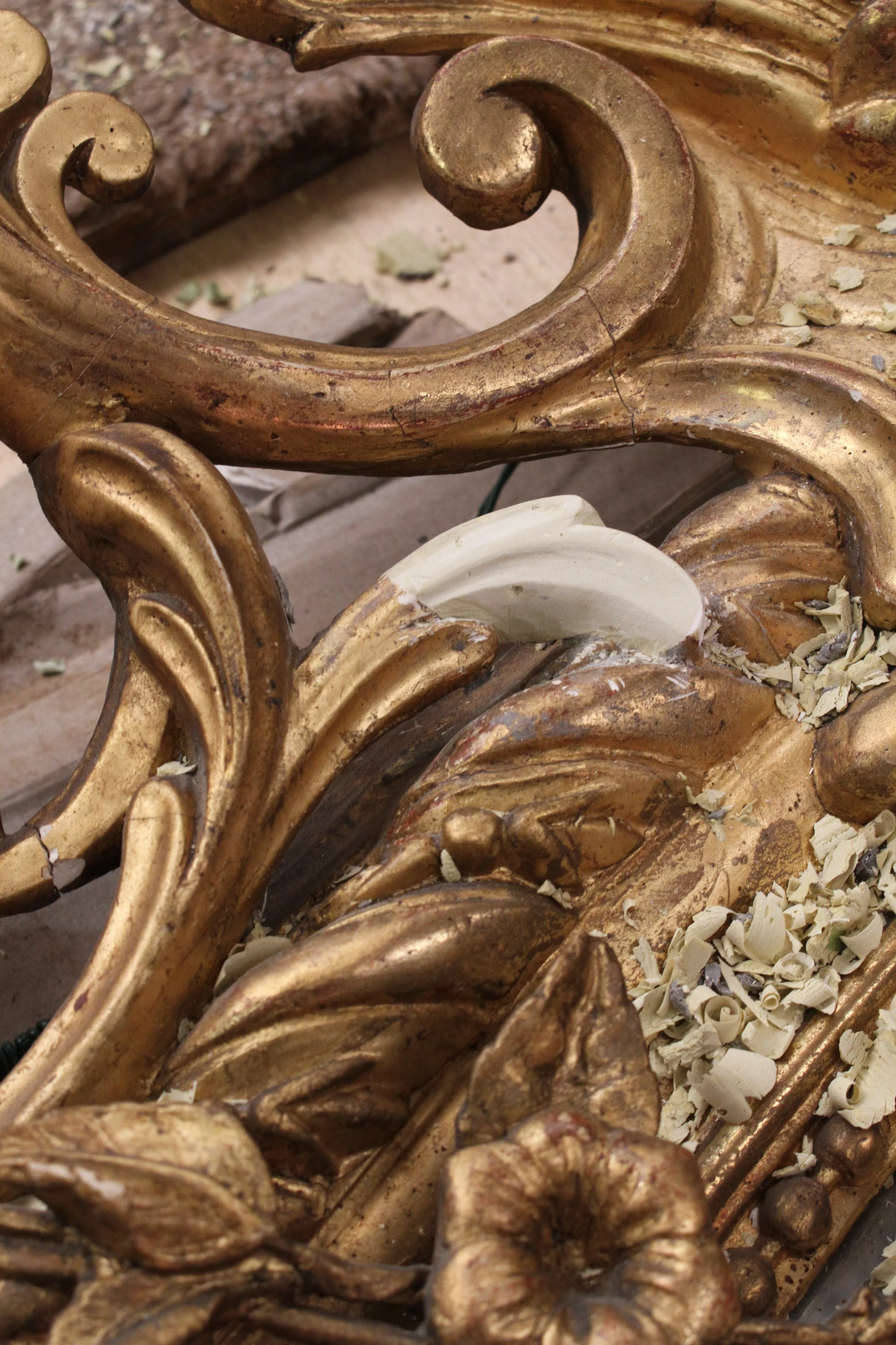 Close-up of an ornate, gold-painted decorative object with intricate scrollwork and floral details, showing signs of aging and chipping paint, with white curled material scattered on its surface.