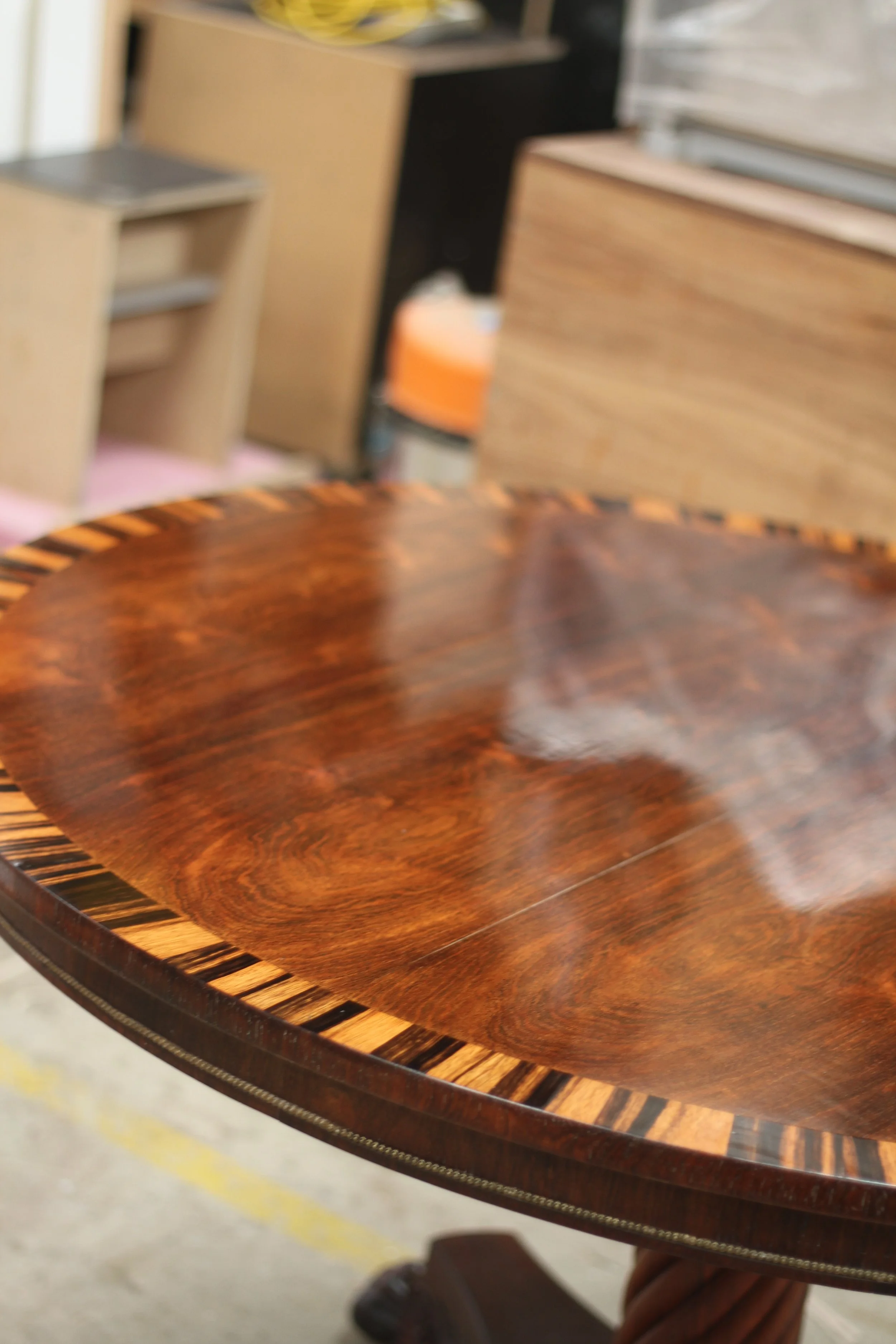 Close-up of a polished wooden round table with a decorative edge, reflecting light, with background showing blurred furniture and concrete floor.