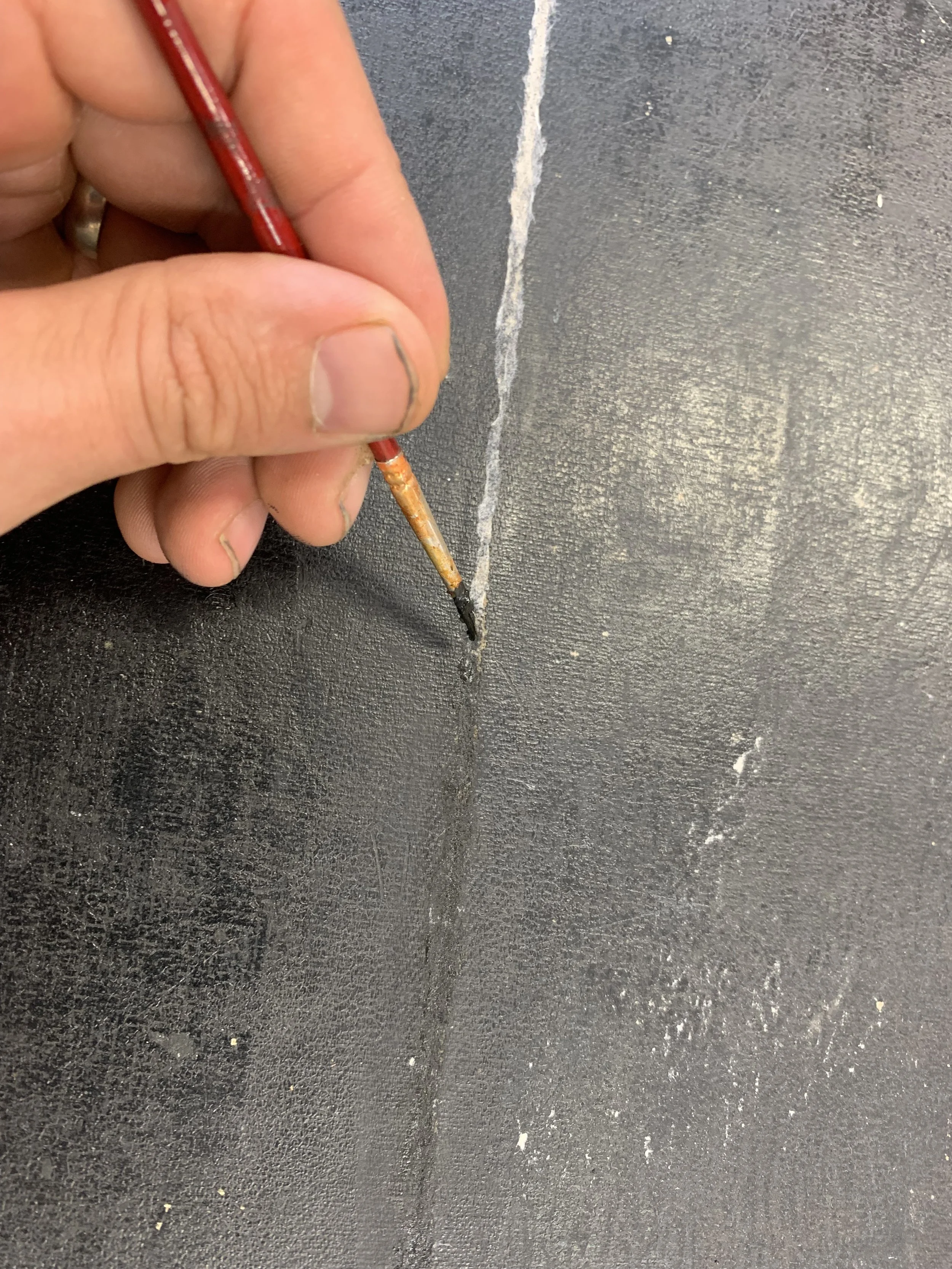 A person holding a small paintbrush and painting over  white line along a feaux leather surface.