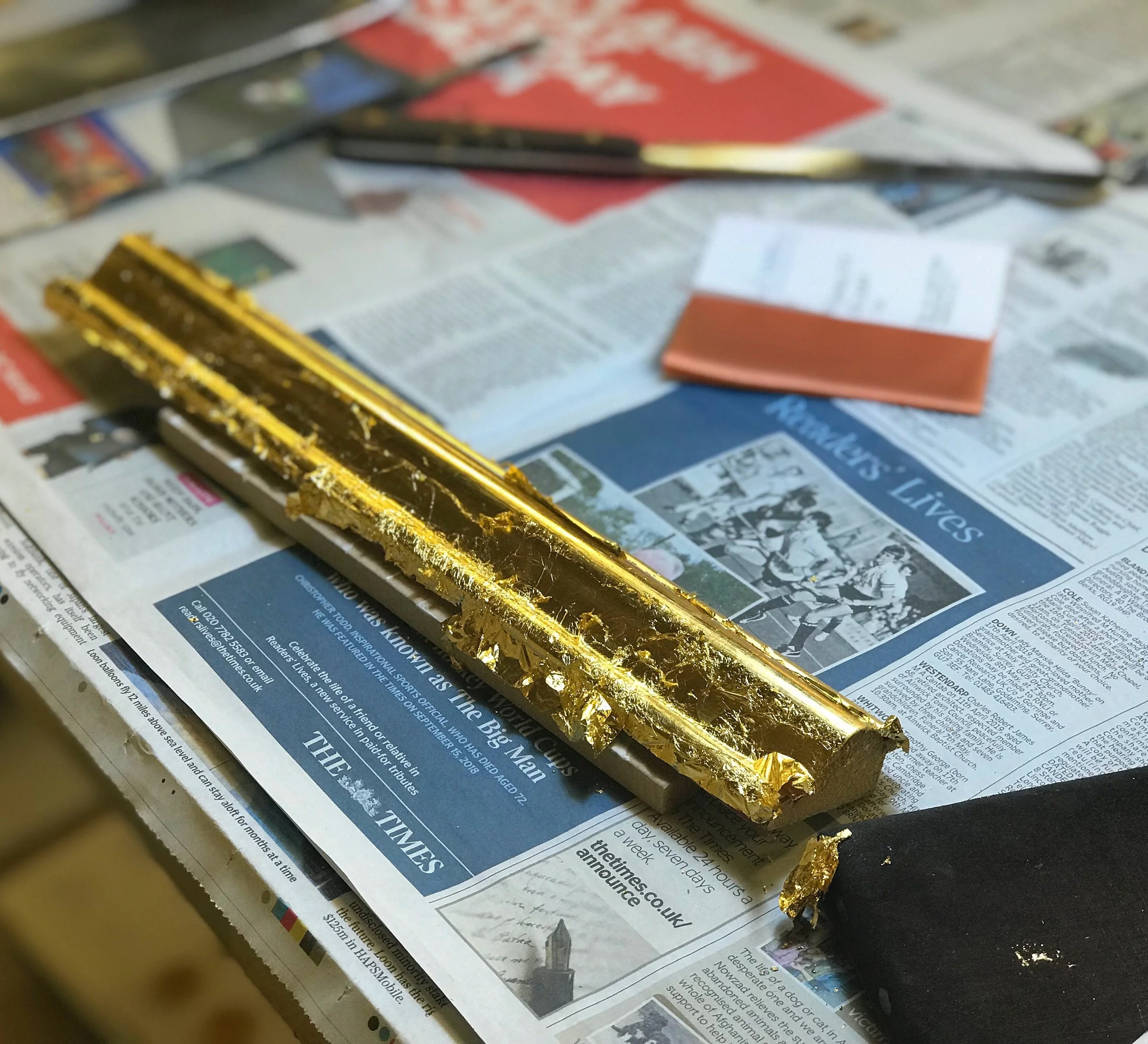 A close-up of a freshly gilt frame moulding on a newspaper, with additional tip and other gilding tools in the background.