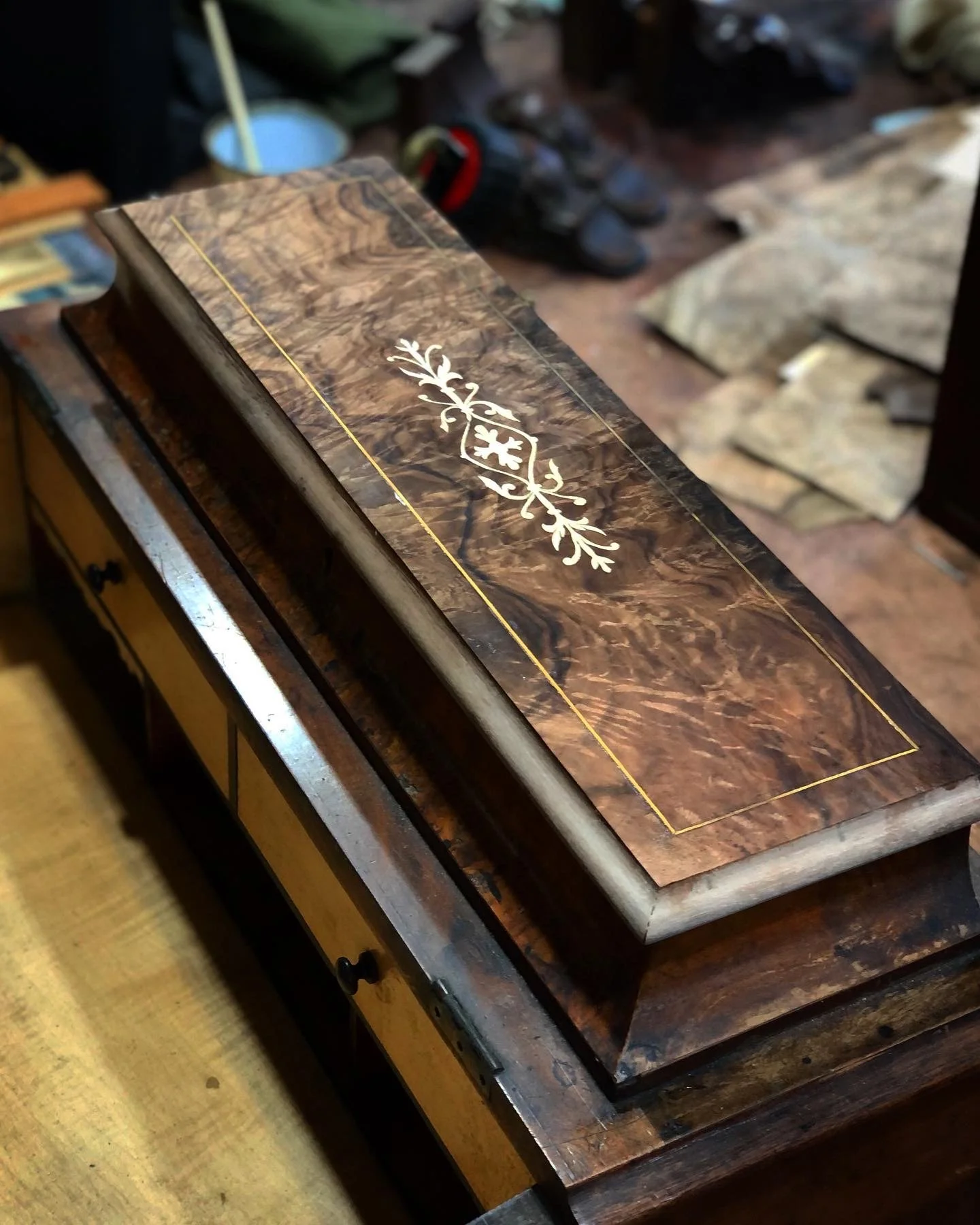 Close-up of a decorative wooden table with intricate floral inlay design in a workshop setting, with woodworking tools and shoes in the background.