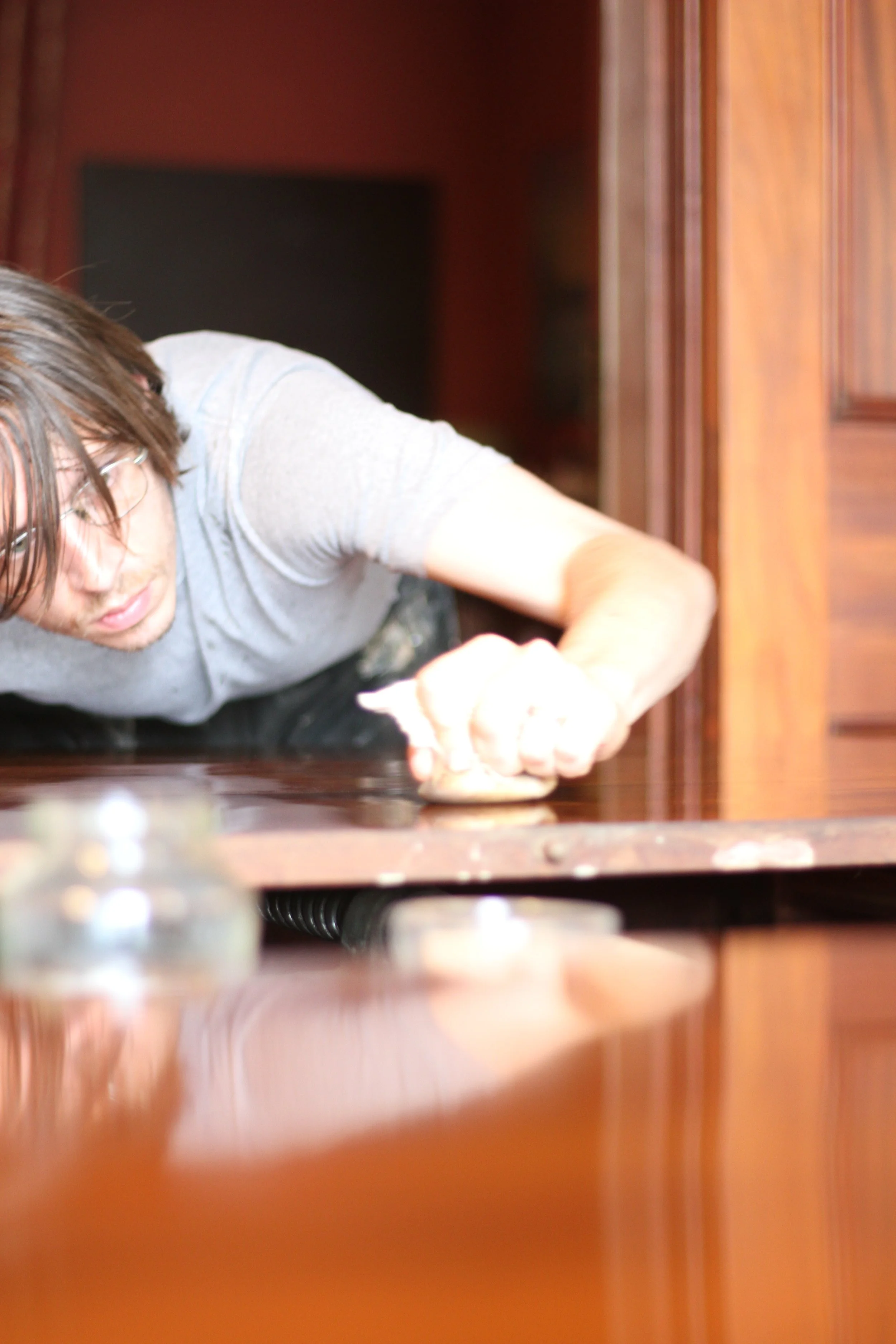 A person with brown hair and glasses is reaching forward on a wooden surface to french polish it, with part of their body visible from an overhead angle. The background shows a wooden paneled wall.